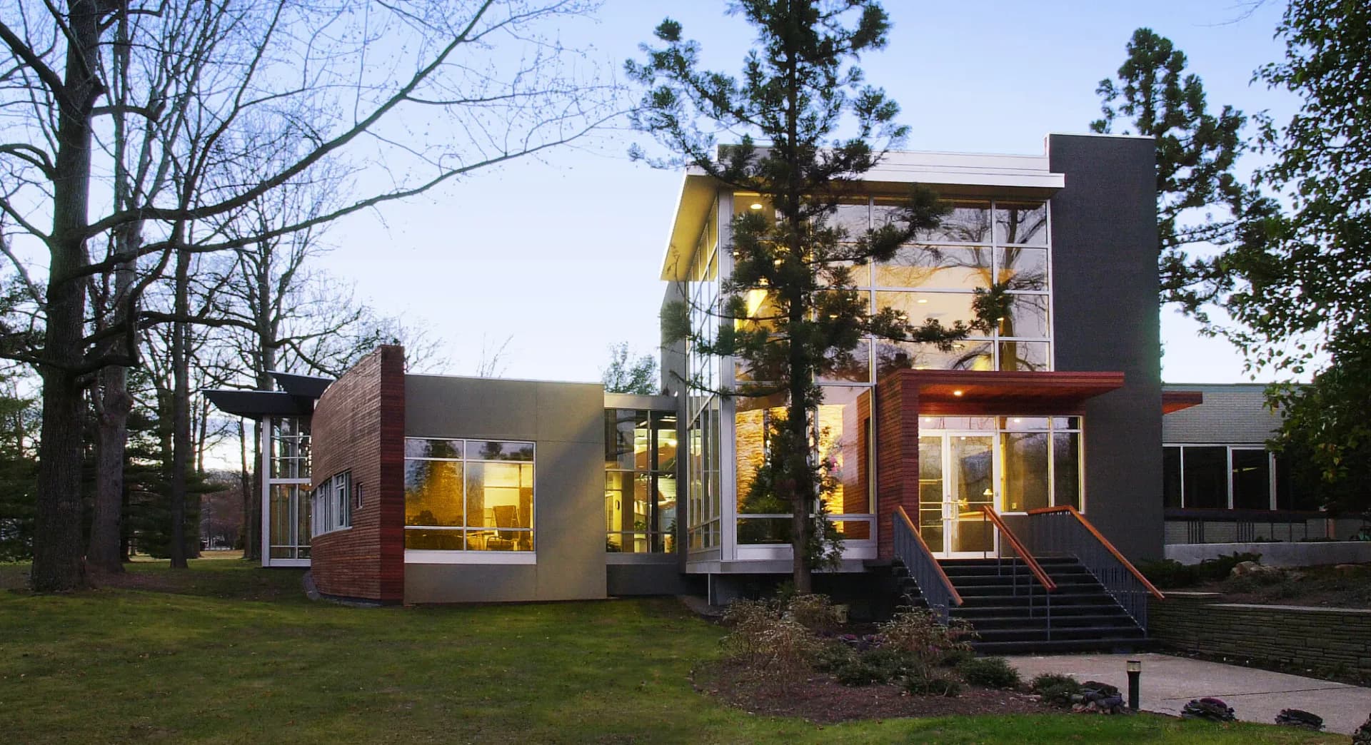 Contemporary residential architecture with mixed materials including brick, steel, and glass volumes, illuminated at dusk, set among mature trees by Terence Smith.