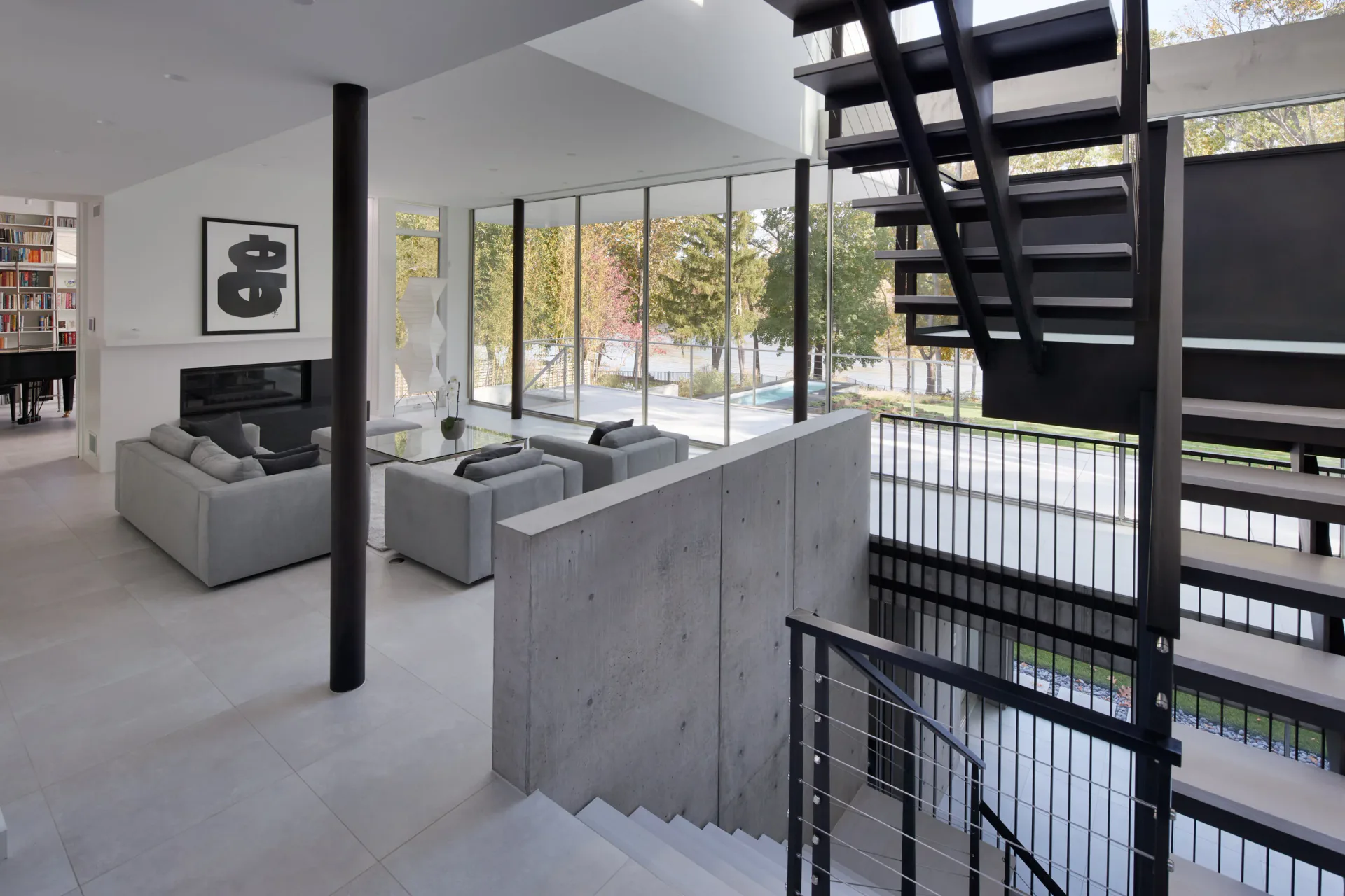Modern loft interior with concrete flooring, black steel staircase and railing, gray seating, large glazed doors overlooking trees, and structural black columns by Terence Smith.
