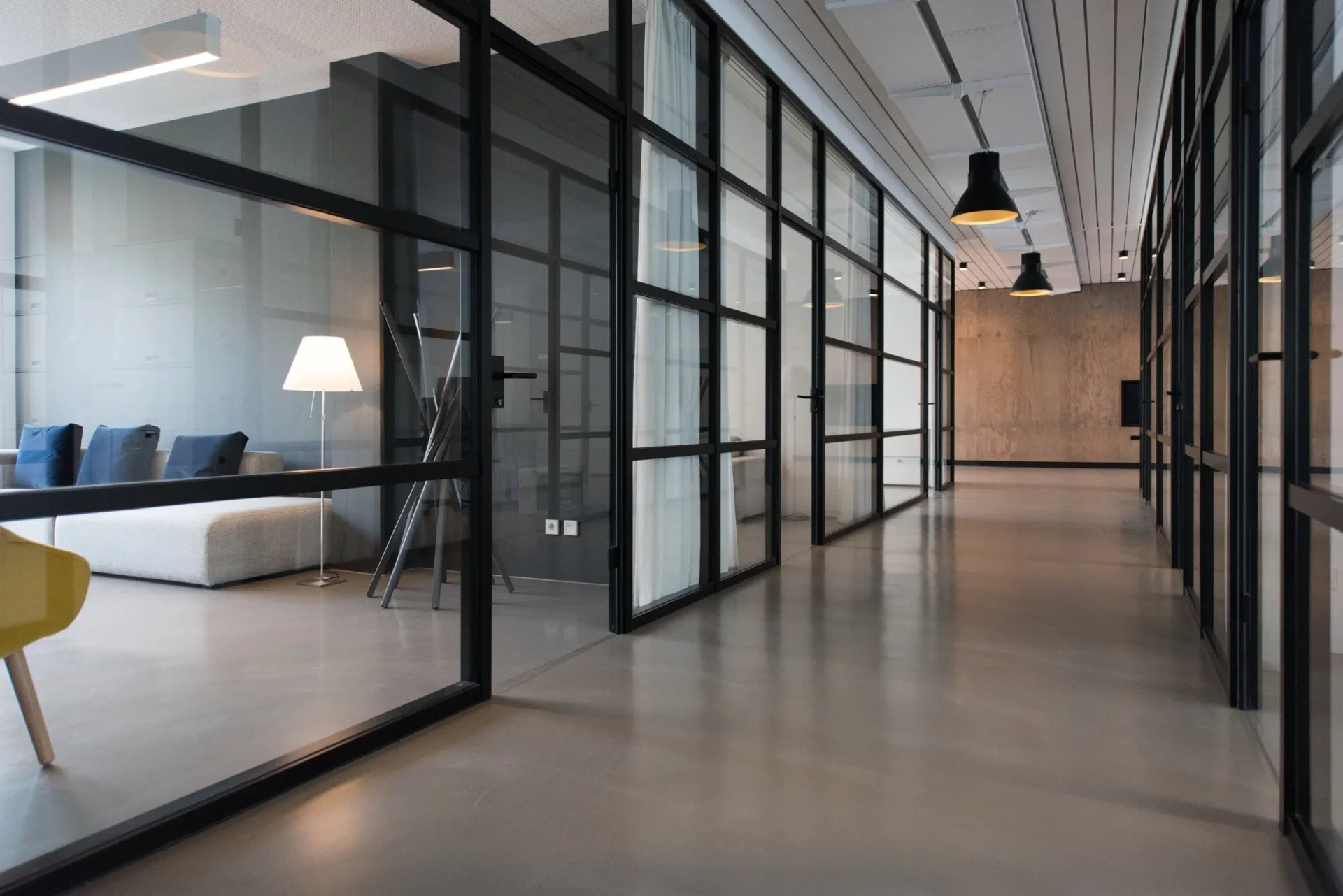 Modern office corridor with glass-partitioned workspaces, polished concrete flooring, and black-framed glass walls by Terry Sachs.