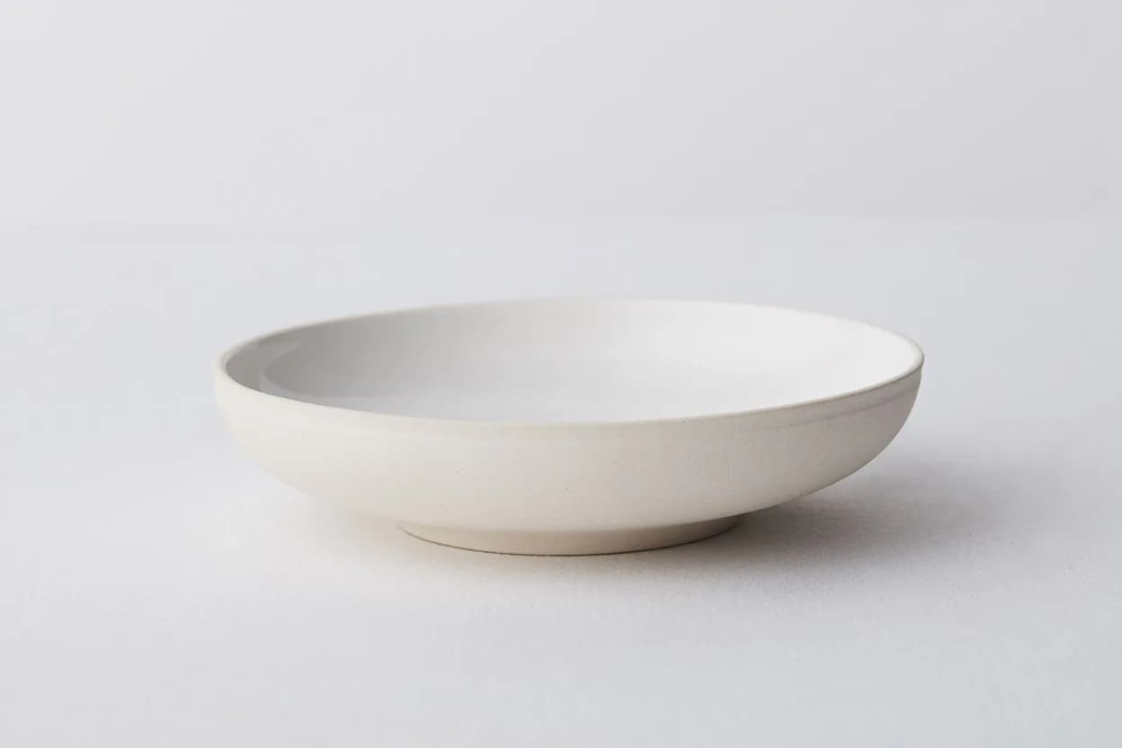 A minimalist white ceramic bowl with a shallow, wide form and soft curved edges rests on a neutral background, showcasing refined ceramic design by Thea von Geldern.