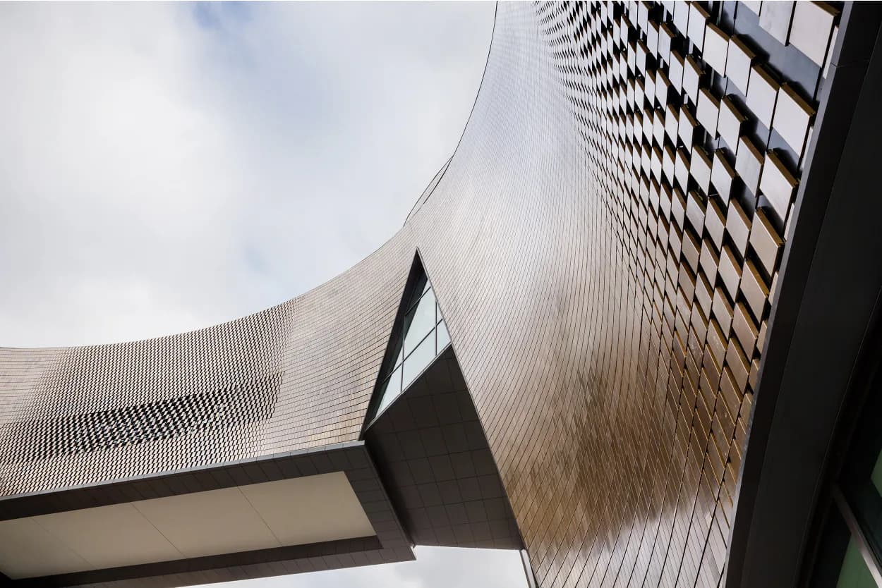 Upward view of curved modernist architecture featuring ribbed metal panels and geometric forms against a cloudy sky, by Thea von Geldern.