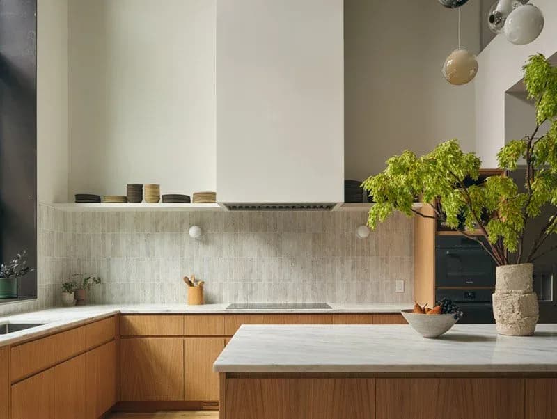 Modern kitchen with warm wood cabinetry, white marble backsplash, concrete countertops, and potted greenery by Themis Haralabides.
