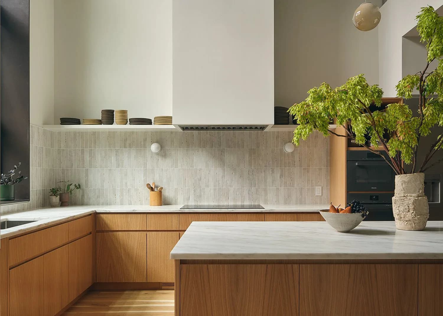 Modern kitchen with warm wood cabinetry, white countertops, vertical tile backsplash, and potted greenery, designed by Themis Haralabides.