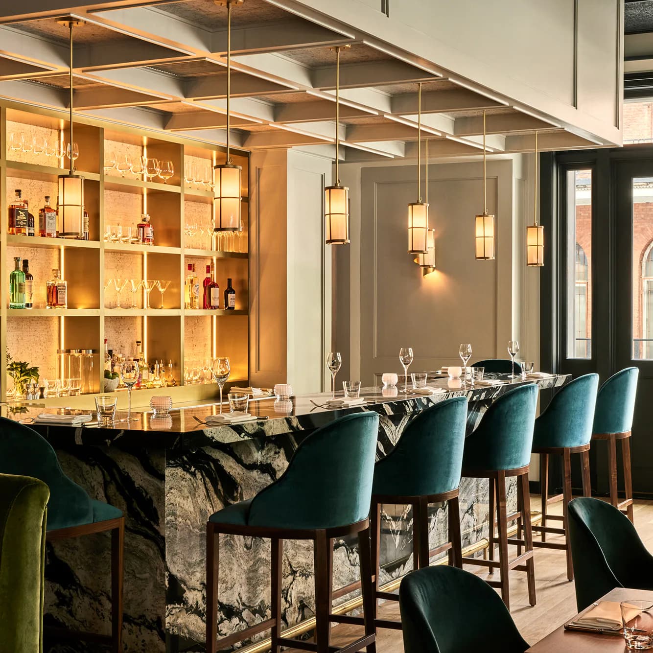 Modern bar interior with teal upholstered stools, dark marble counter, brass-framed glass partition, and illuminated shelving displaying spirits and glassware by Theo Guo.