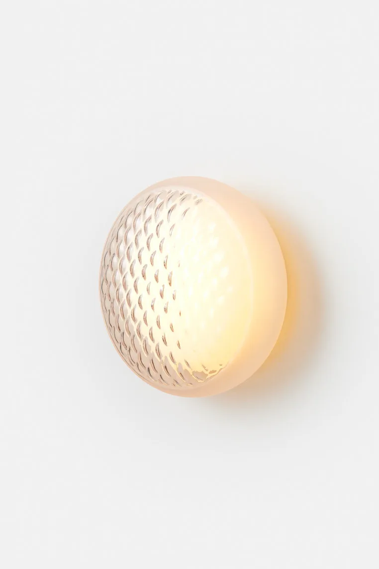 Cream-colored spherical wall sconce with textured mesh pattern on left side, emitting warm light against white wall by Theo Richardson.