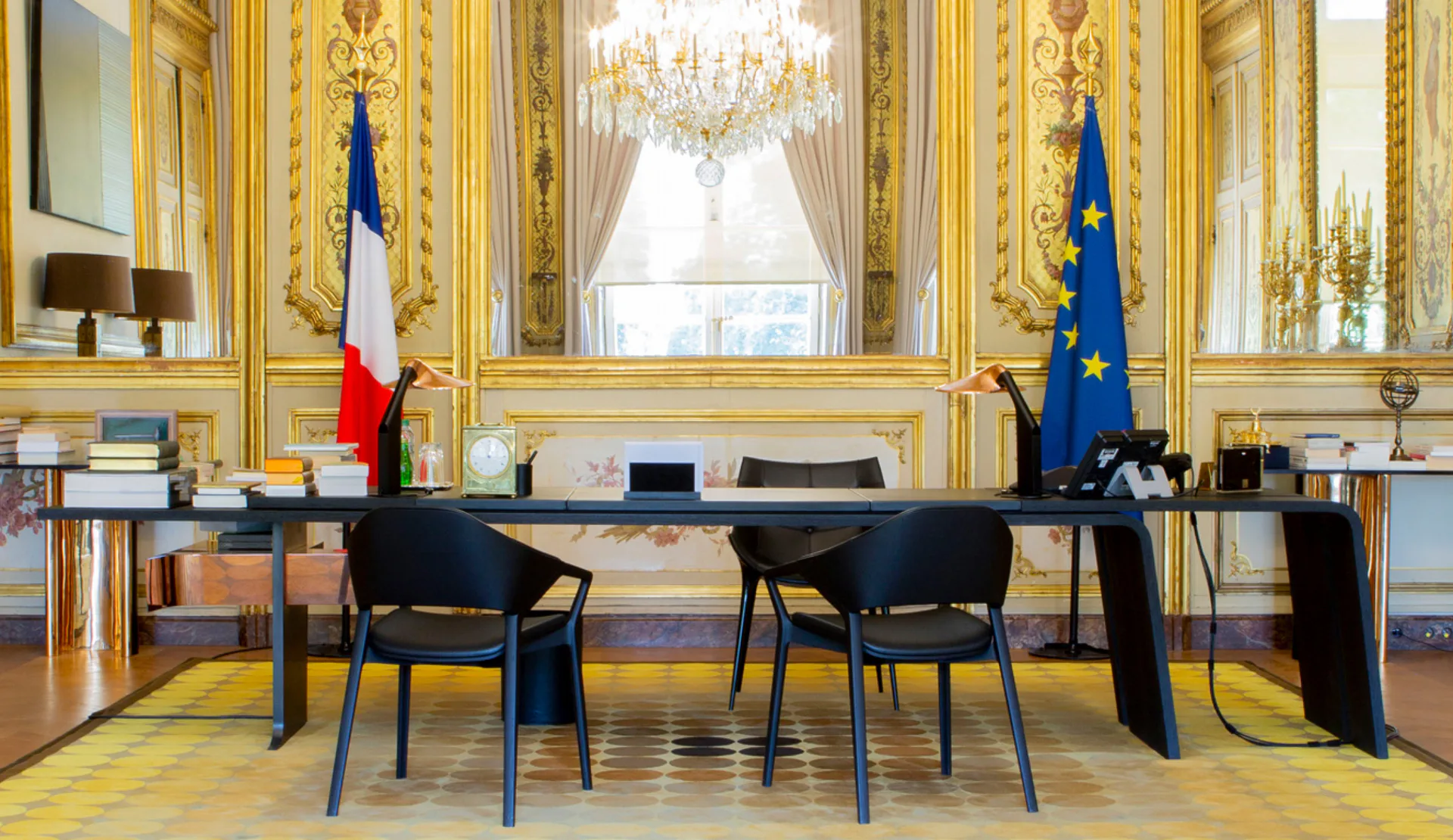 Elegant formal office with gilded paneling, crystal chandelier, and French flags, featuring a minimalist black desk and chairs by Thierry Lemaire.