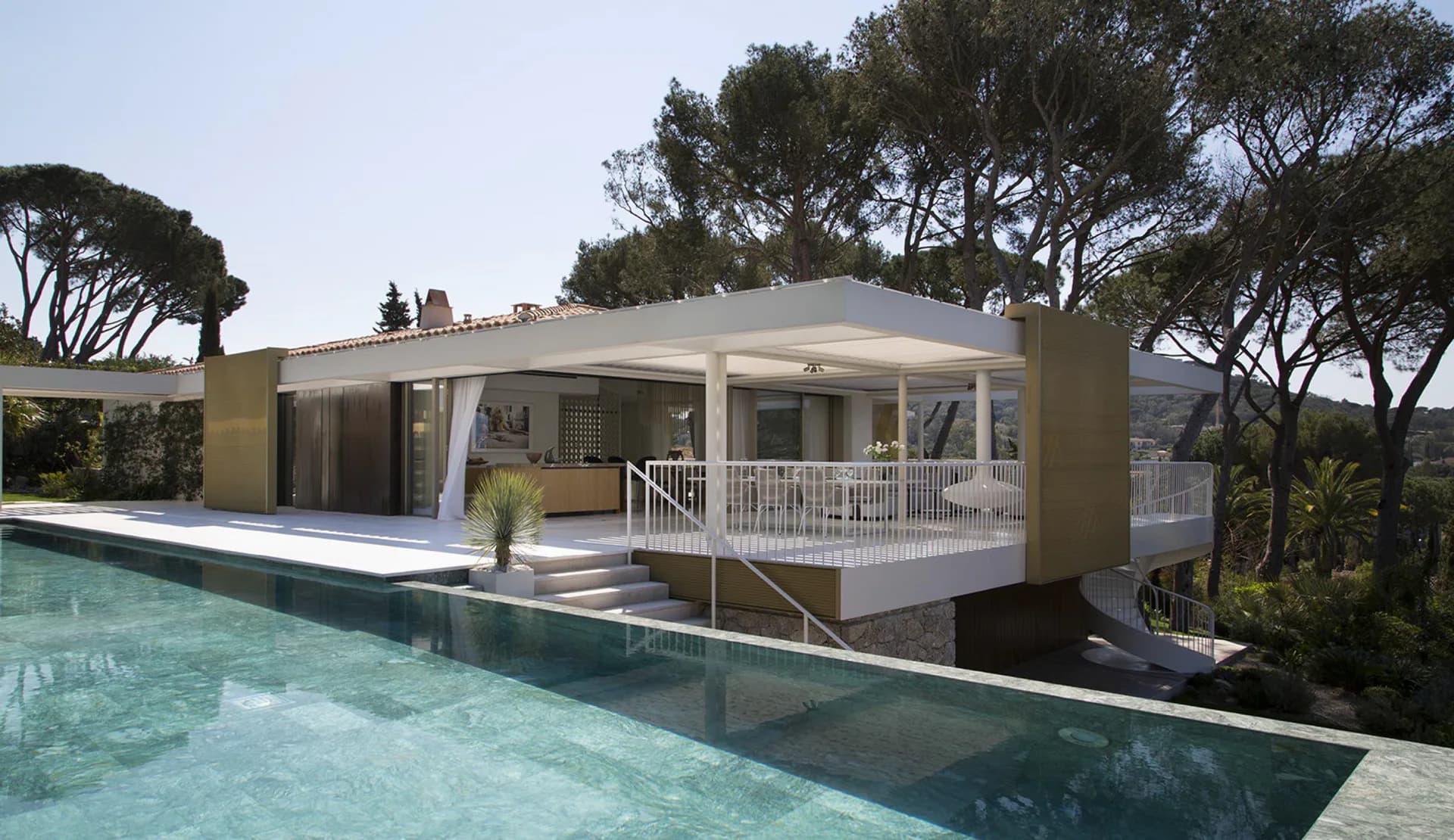 Modern residential villa with clean-lined architecture, cream and tan exterior, white railings, and large glazed openings overlooking an infinity pool in a Mediterranean landscape setting, designed by Thierry Lemaire.