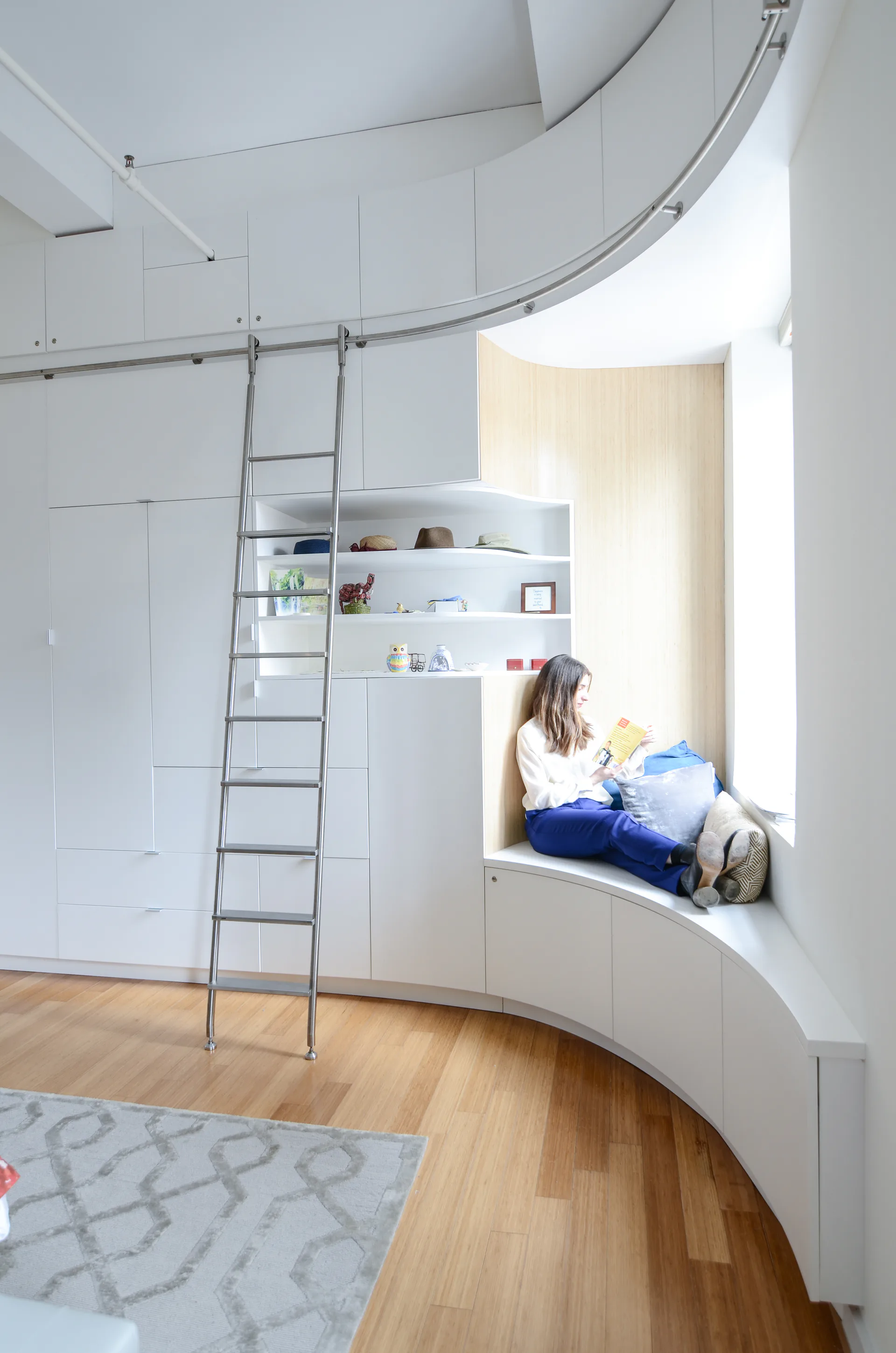Modern bedroom with curved white built-in cabinetry, rolling library ladder, wooden flooring, and a bright window nook by Thomas Diorio.