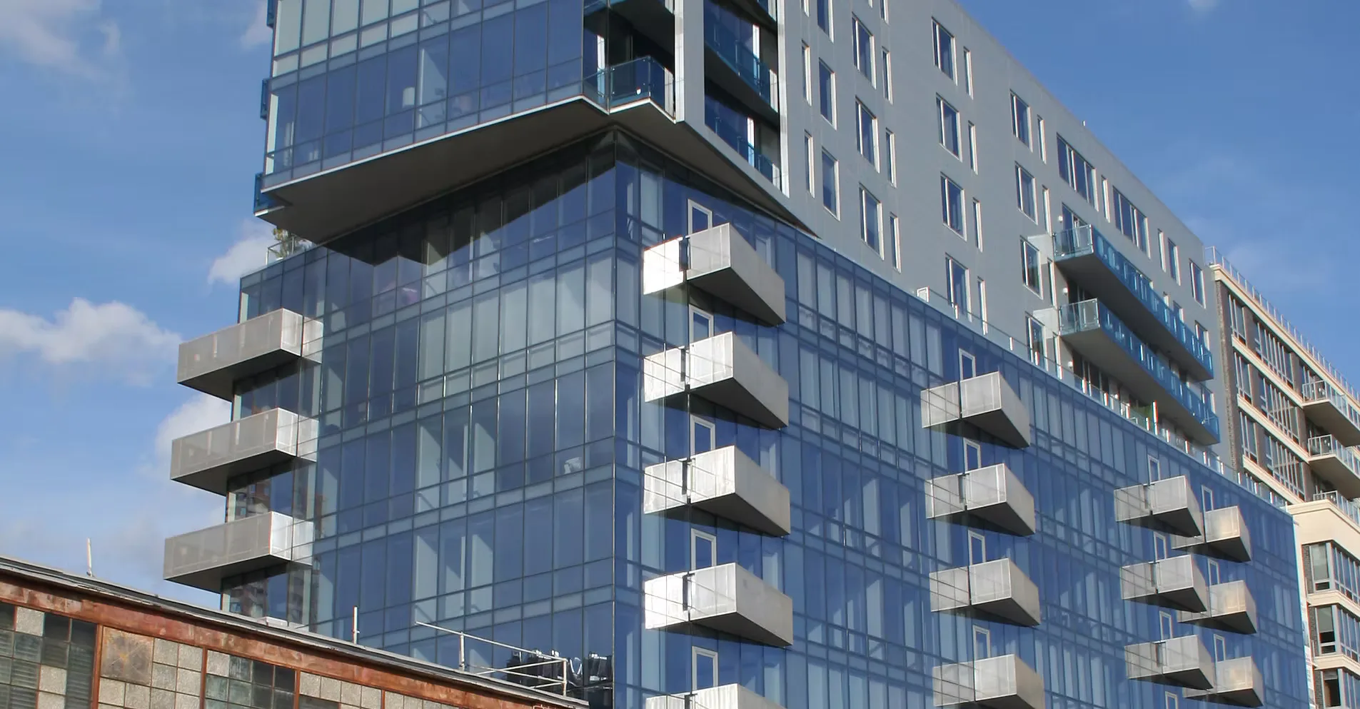 Modern residential building with glass and metal facade featuring stacked balconies and mixed materials, bright daylight by Thomas Gilman.