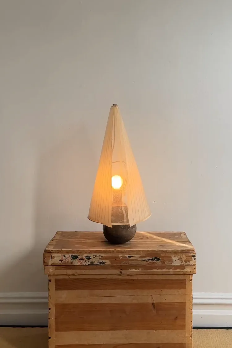 Wooden crate base supporting a cone-shaped pendant lamp with warm amber fabric shade and illuminated interior, demonstrating minimalist design by Thomas Yang.