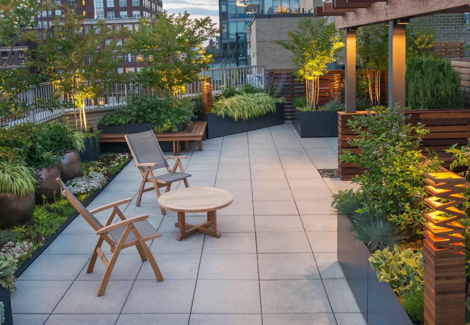 Modern rooftop terrace with light gray paving, wooden seating, lush greenery, and warm uplighting by Tillie O'Reilly.