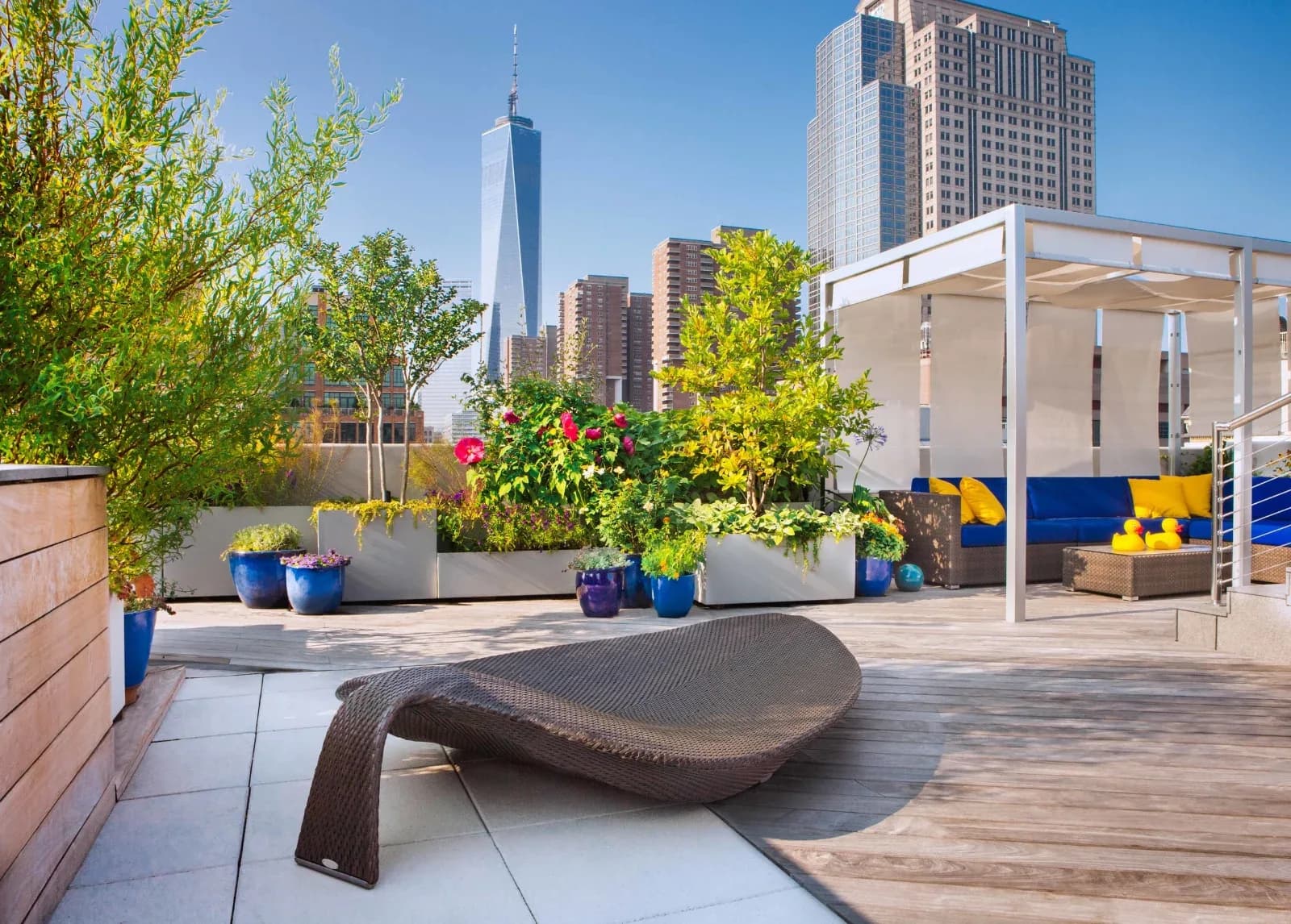 A rooftop terrace with modern sculptural seating, blue planters, lush greenery, and Manhattan skyline views, designed by Tillie O'Reilly.