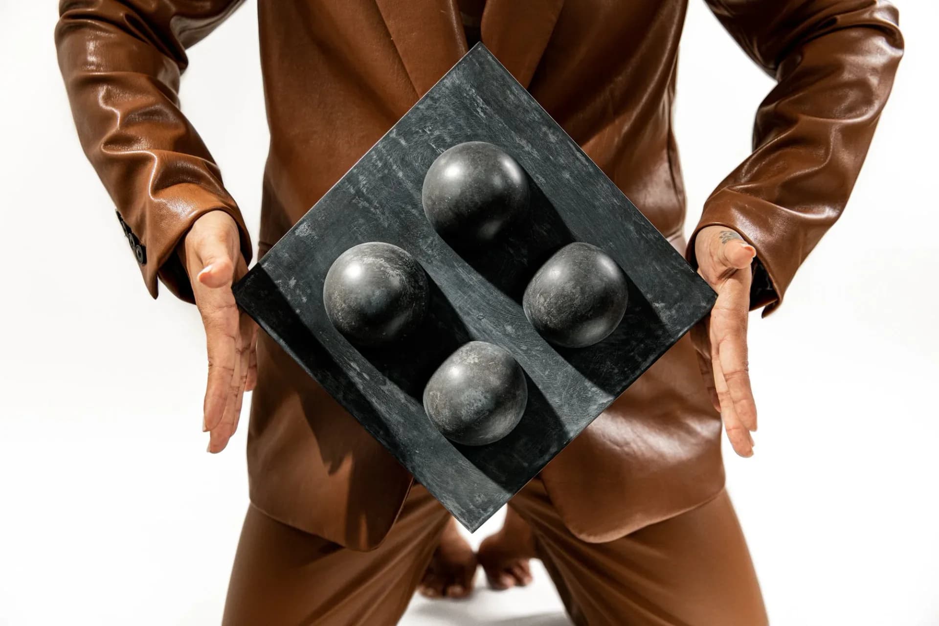 Man in brown leather jacket holding a dark stone or concrete panel with four spherical metal balls embedded in it, showcasing a sculptural product design by Tina Scepanovic.