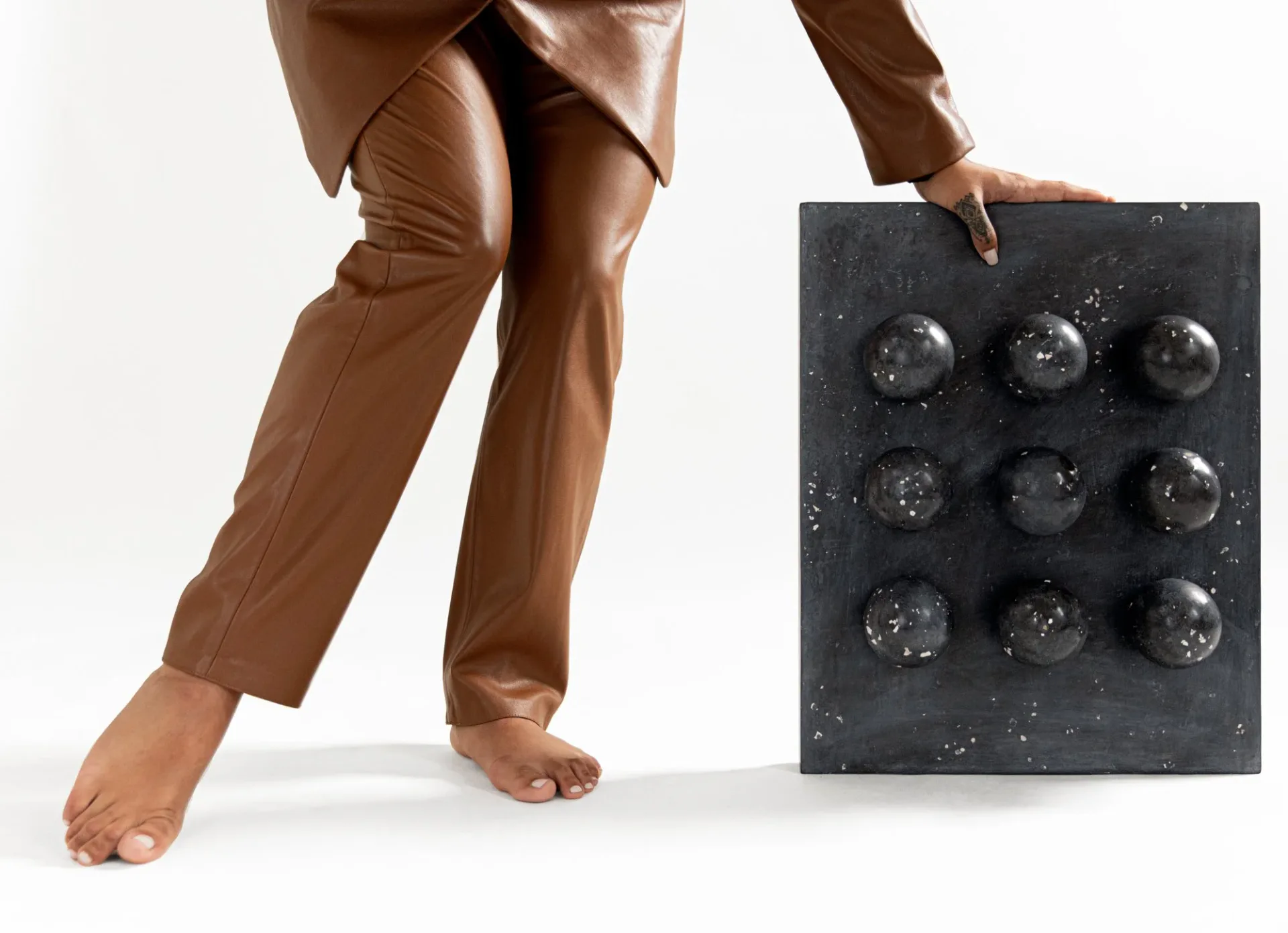 Tactile installation featuring a dark textured panel with nine spherical elements arranged in a grid, complemented by draped bronze fabric and barefoot figure, by Tina Scepanovic.