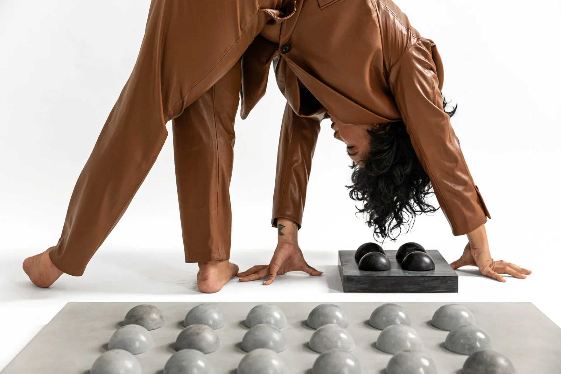 Abstract sculptural installation featuring a person in a brown outfit bending over white geometric forms arranged on a platform, exploring spatial dynamics and material contrast by Tina Scepanovic.