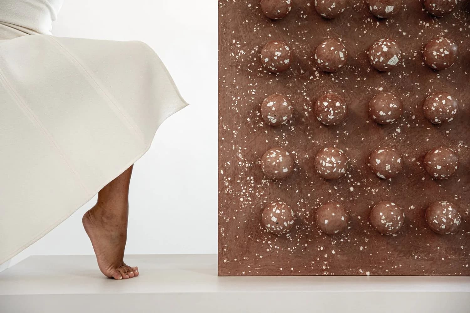Minimalist interior installation featuring a draped white fabric element and textured brown wall panel with spherical relief details, by Tina Scepanovic.