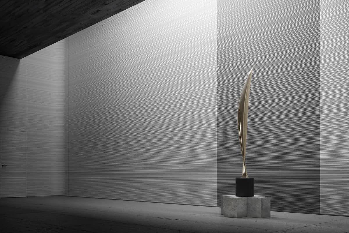 Minimalist interior space with textured white walls, concrete floor, and a sculptural brass vessel on a stone pedestal by Todd Bracher.
