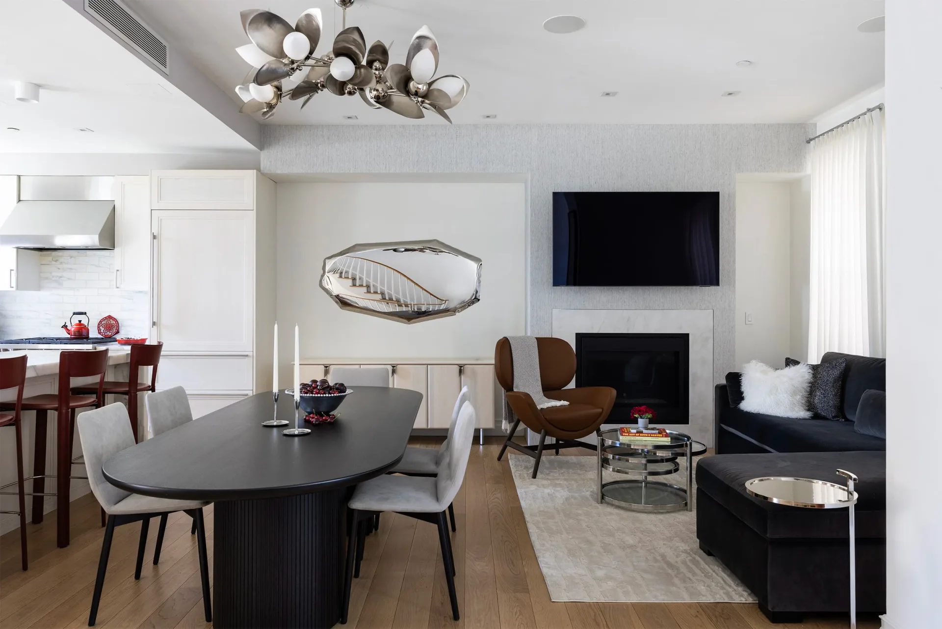Modern open-plan living and dining space featuring a black round dining table, fireplace with TV, dark sofa, and brass chandelier by Tolu Odunfa Dragone.