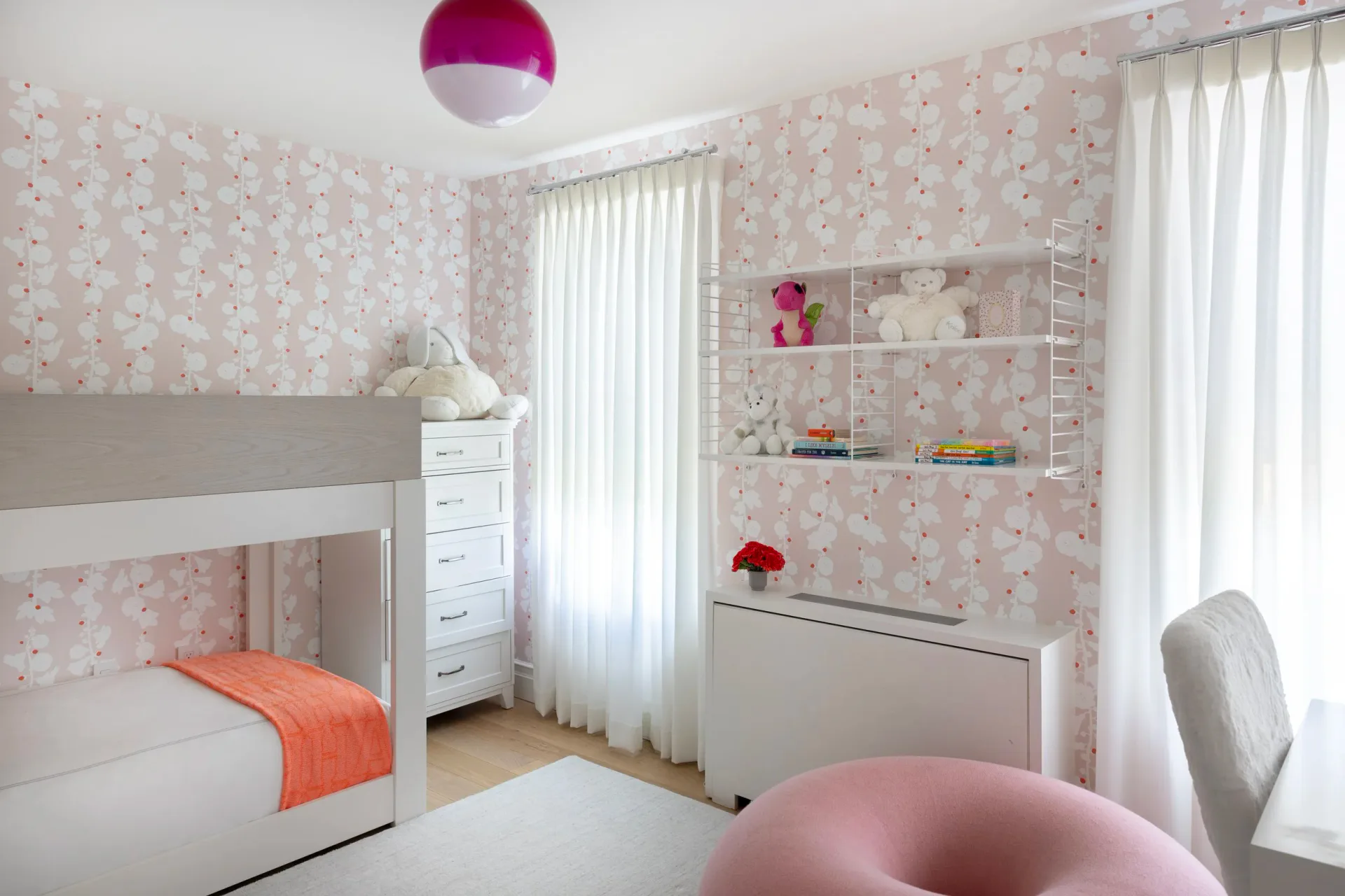 A contemporary girl's bedroom with soft pink patterned wallpaper, white furniture, built-in shelving, and bright natural light from sheer curtains, designed by Tolu Odunfa Dragone.