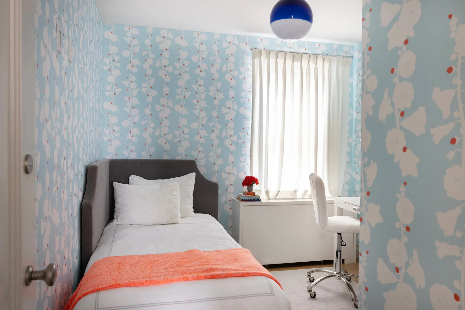 A bright child's bedroom features blue patterned wallpaper, a charcoal upholstered bed with white bedding and coral throw, white desk, and natural light from curtained window, designed by Tolu Odunfa Dragone.