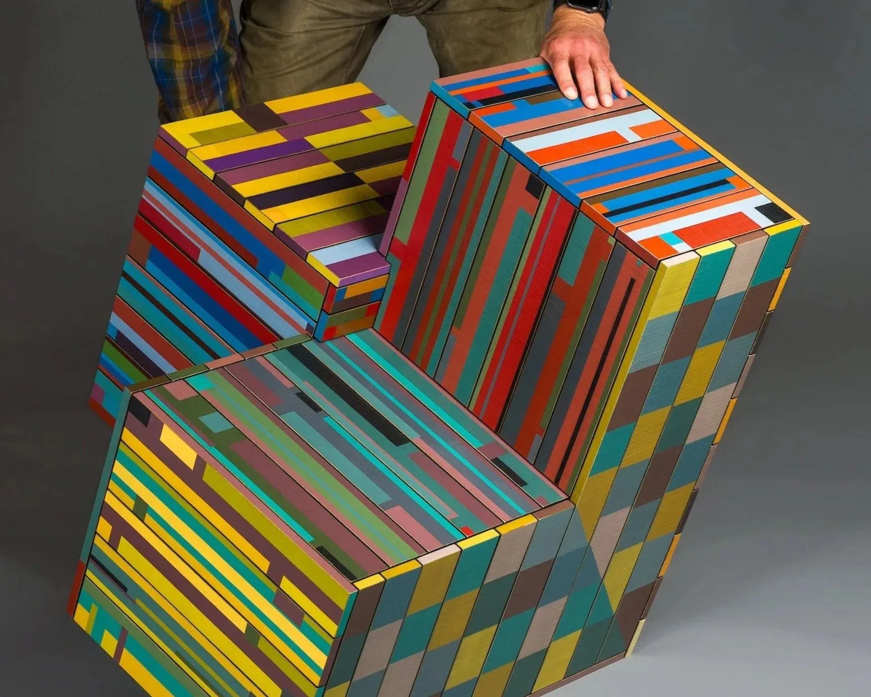 Colorful striped wooden storage boxes with geometric patterns in blue, red, yellow, and green, displayed as a modular furniture installation by Tom Loeser.