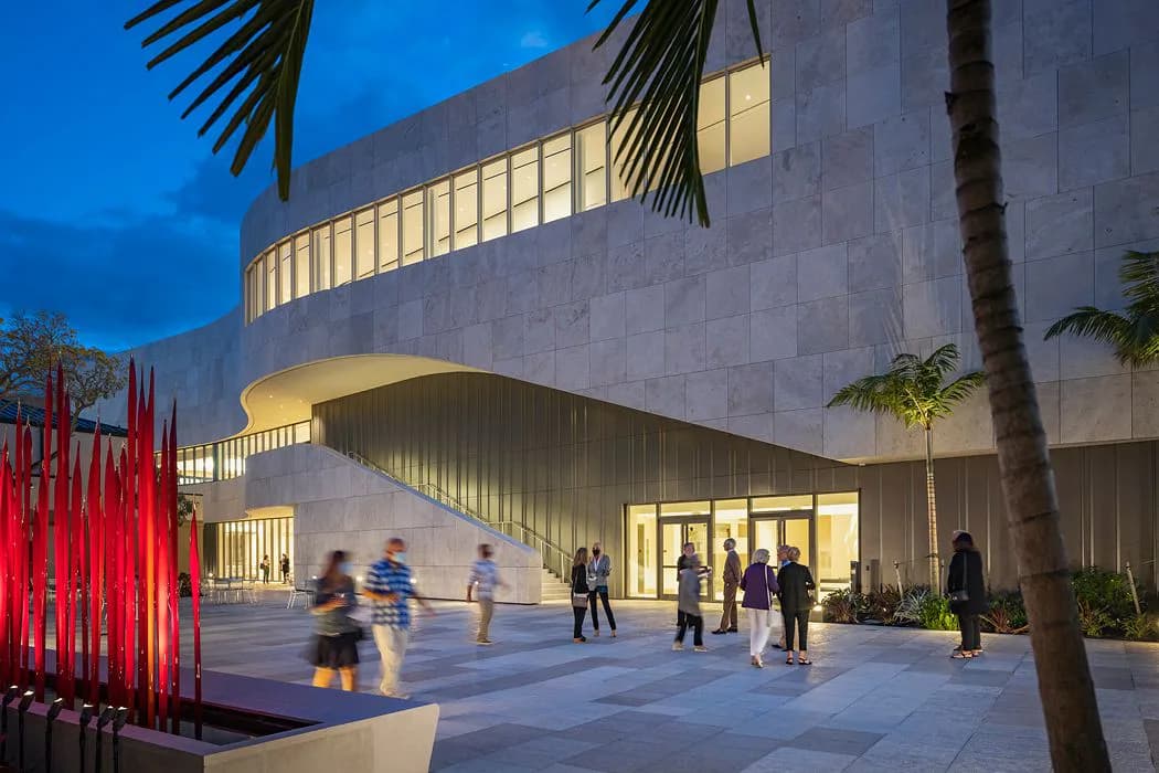 Modern architectural rendering of a curved concrete building with illuminated window bands at dusk, featuring a plaza with red sculptural elements and palm trees, designed by Tomoko Akiba.