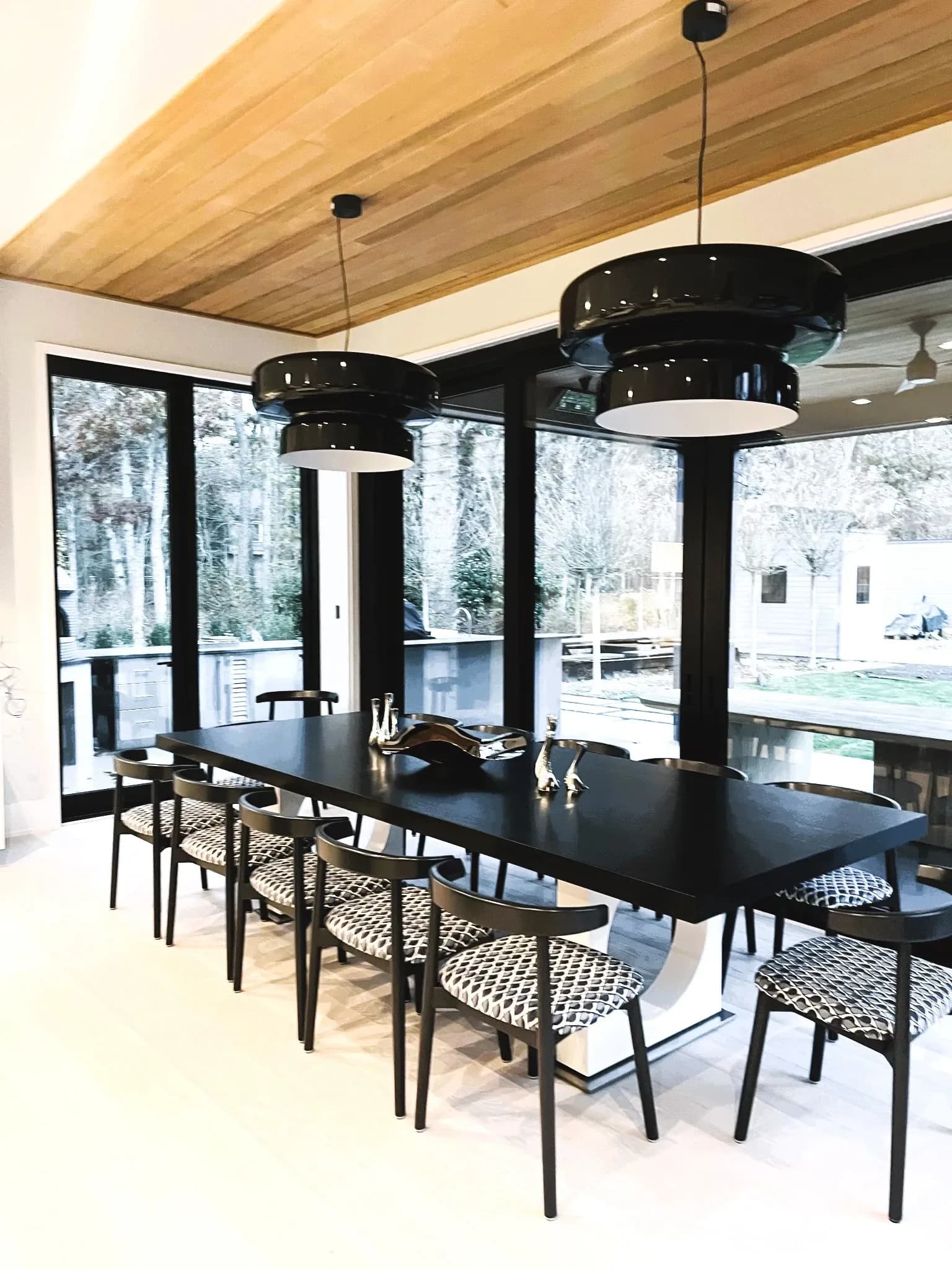 Modern dining room with black rectangular table, woven chairs, two black pendant lights, wood ceiling, and floor-to-ceiling windows overlooking outdoor space by Tony Klein.