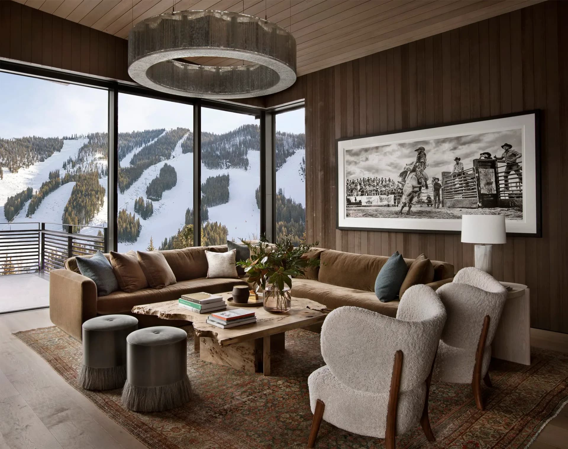 Luxury alpine lounge with brown wood walls, curved sectional sofa, modern ceiling fixture, and expansive mountain views through floor-to-ceiling windows by Tori Catherall.