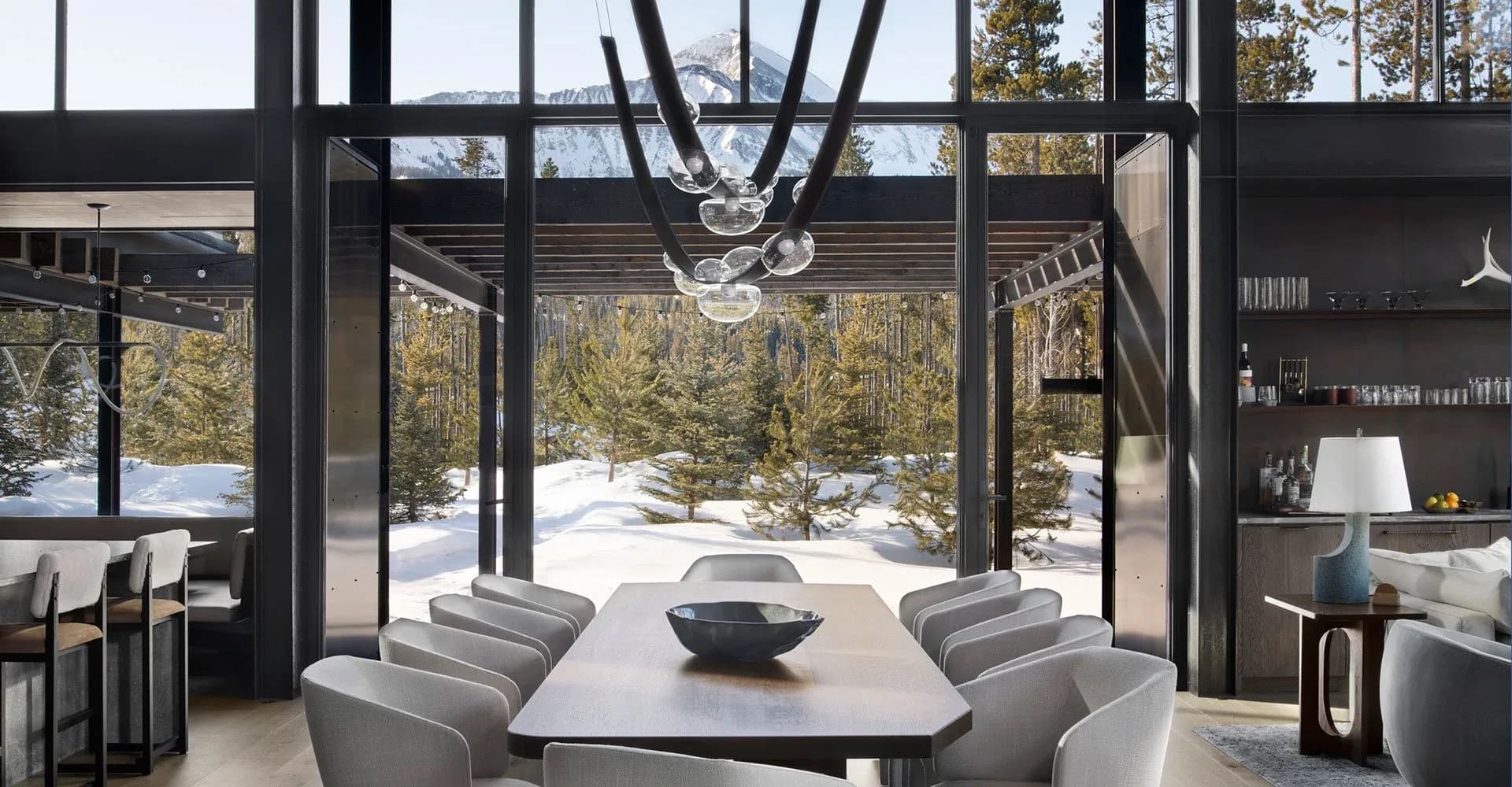 Modern dining space with black steel framing, gray upholstered chairs, white table, and expansive windows overlooking snow-covered landscape by Tori Catherall.