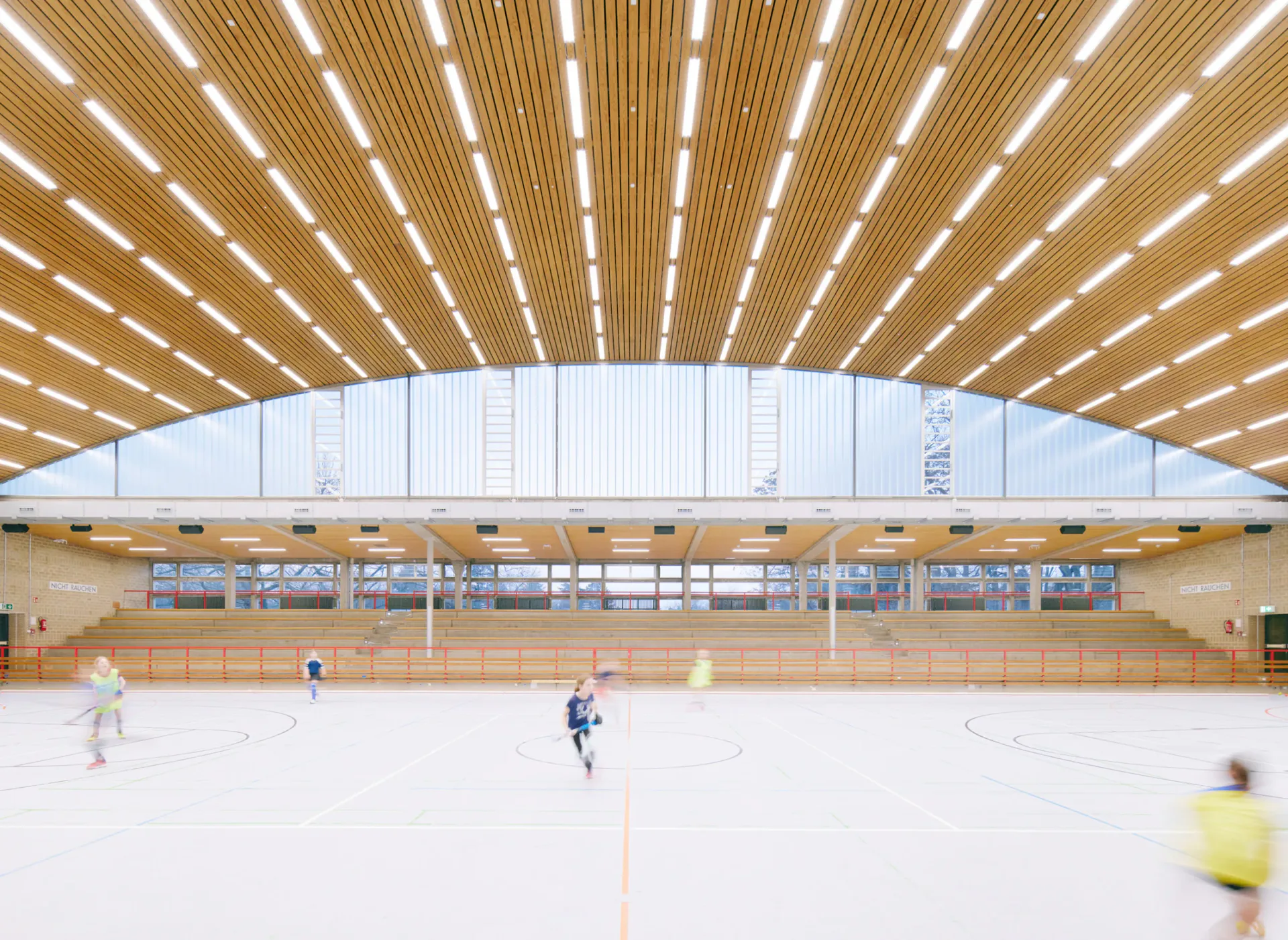 Bright modern sports hall with radiating wooden ceiling slats, white walls, and indoor court with players, designed by Trevor Wood.
