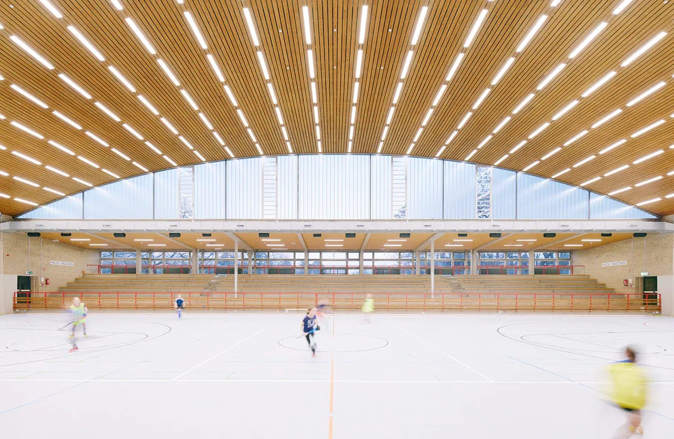 Modern sports hall interior with curved wooden ceiling slats, white walls, and indoor court with players, designed by Trevor Wood.