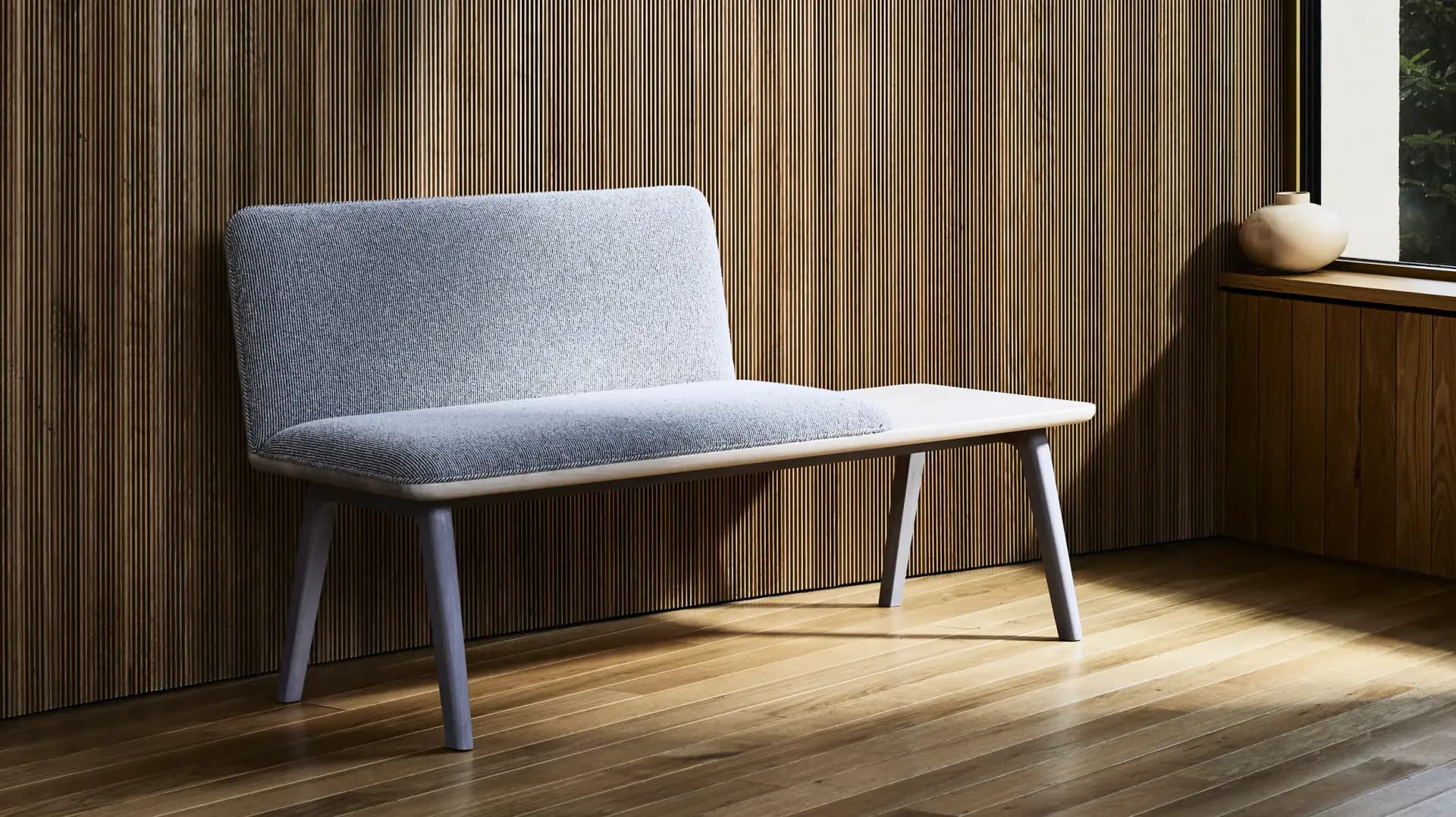 Modern minimalist interior with a light blue upholstered bench against vertical wood paneling, warm wooden flooring, and natural light, designed by Tugba Ozkan.