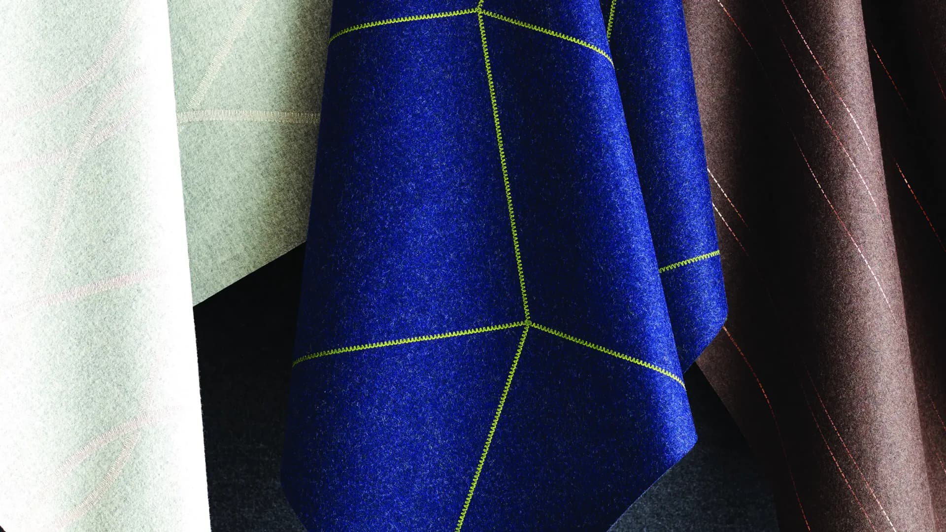 Close-up of folded blue wool textile with yellow topstitching detail, displayed alongside neutral and brown fabric samples by Tugba Ozkan.