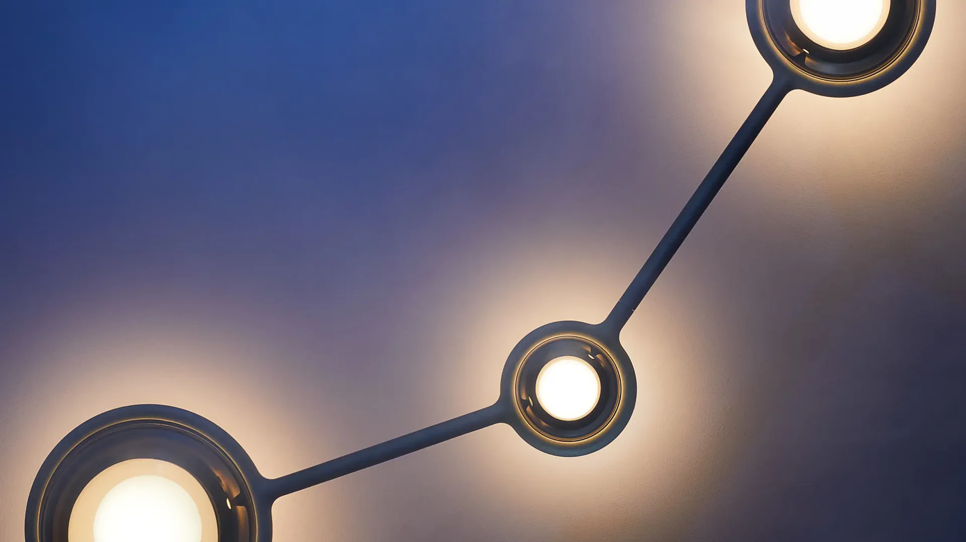 Contemporary pendant lighting installation with three geometric fixtures connected by curved arms against a soft gradient background by Tugba Ozkan.