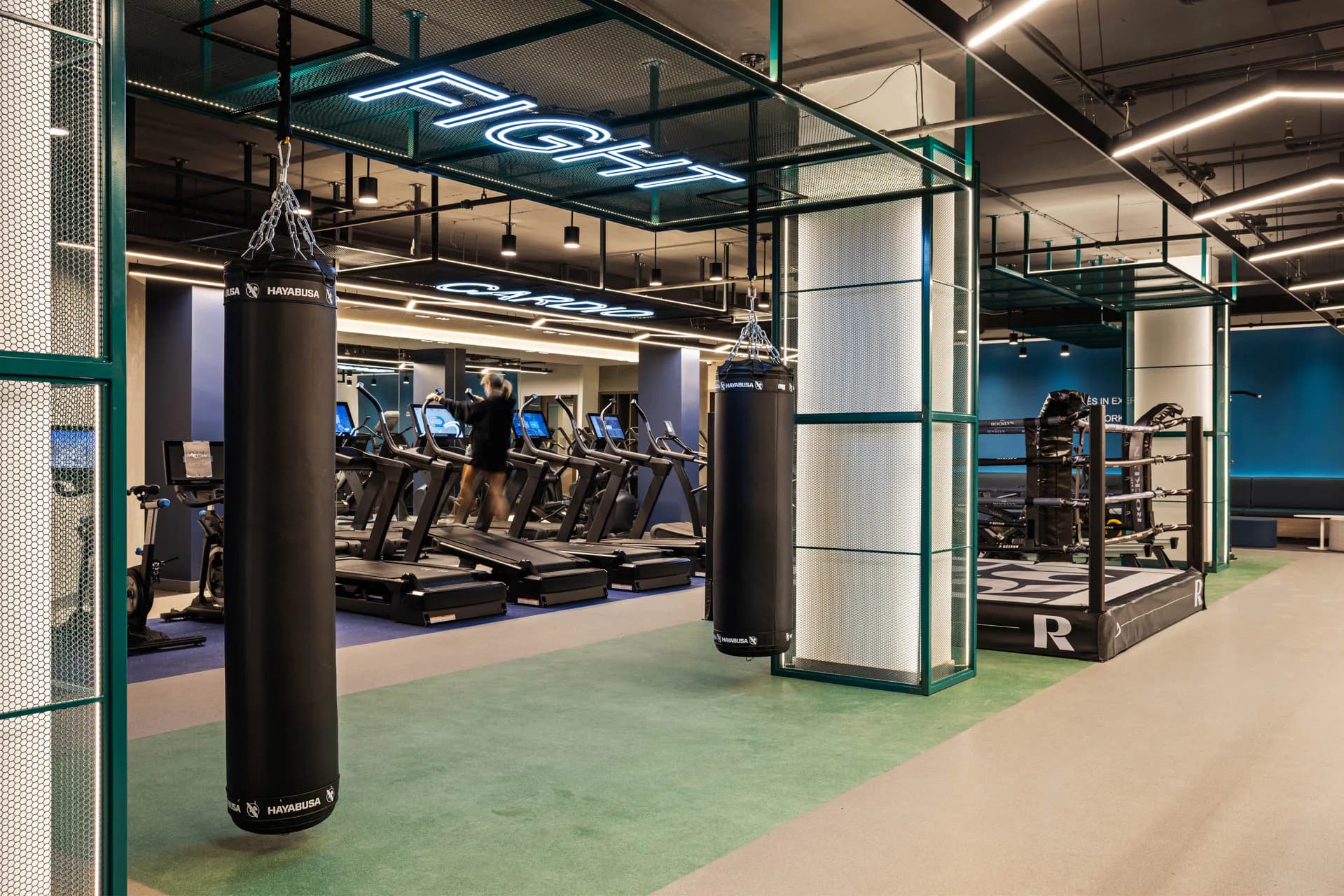 Modern gym interior with green flooring, black support columns, cardio equipment visible behind glass partitions, and blue neon signage by Tugce Kodalak.