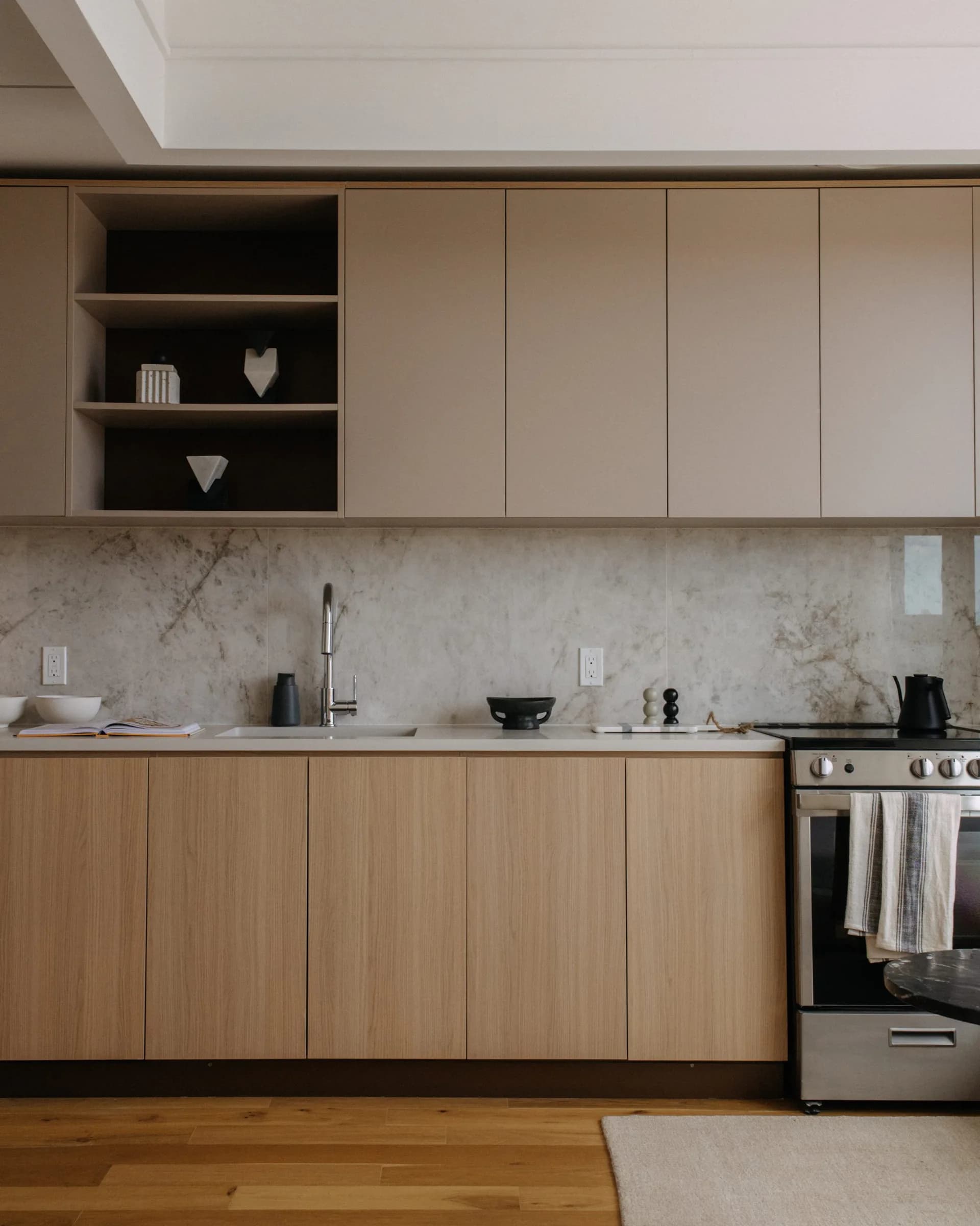 Modern kitchen with warm wood lower cabinetry, neutral upper cabinets, marble backsplash, stainless steel appliances, and open shelving with black backing by Tugce Kodalak.
