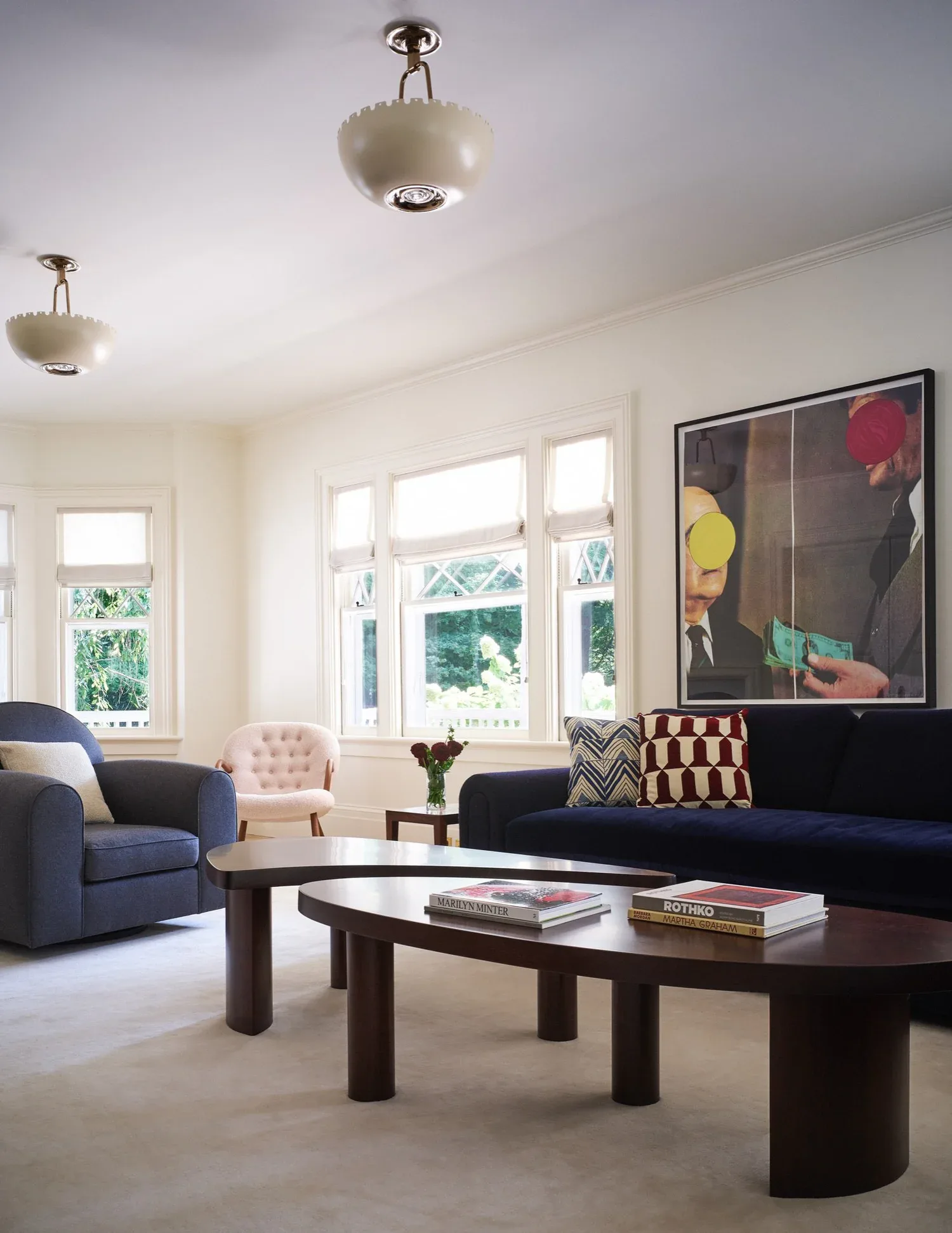 Modern living room with blue armchair, black sofa, dark wood oval coffee table, large windows, pendant lighting, and colorful abstract artwork by Tusy Culos.