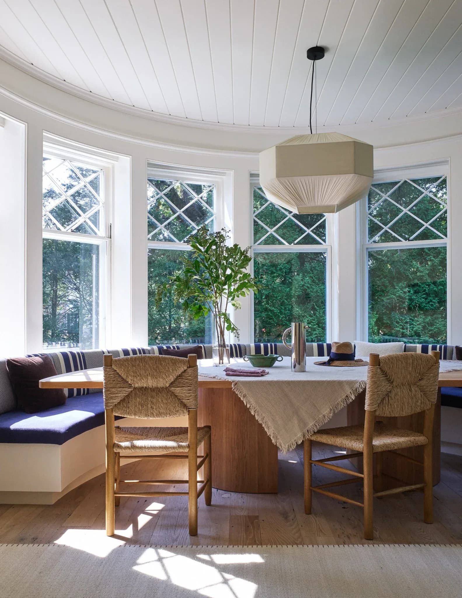 Bright sunroom with curved walls, large windows overlooking trees, wooden dining table with woven chairs, striped banquette seating, and pendant lighting by Tusy Culos.