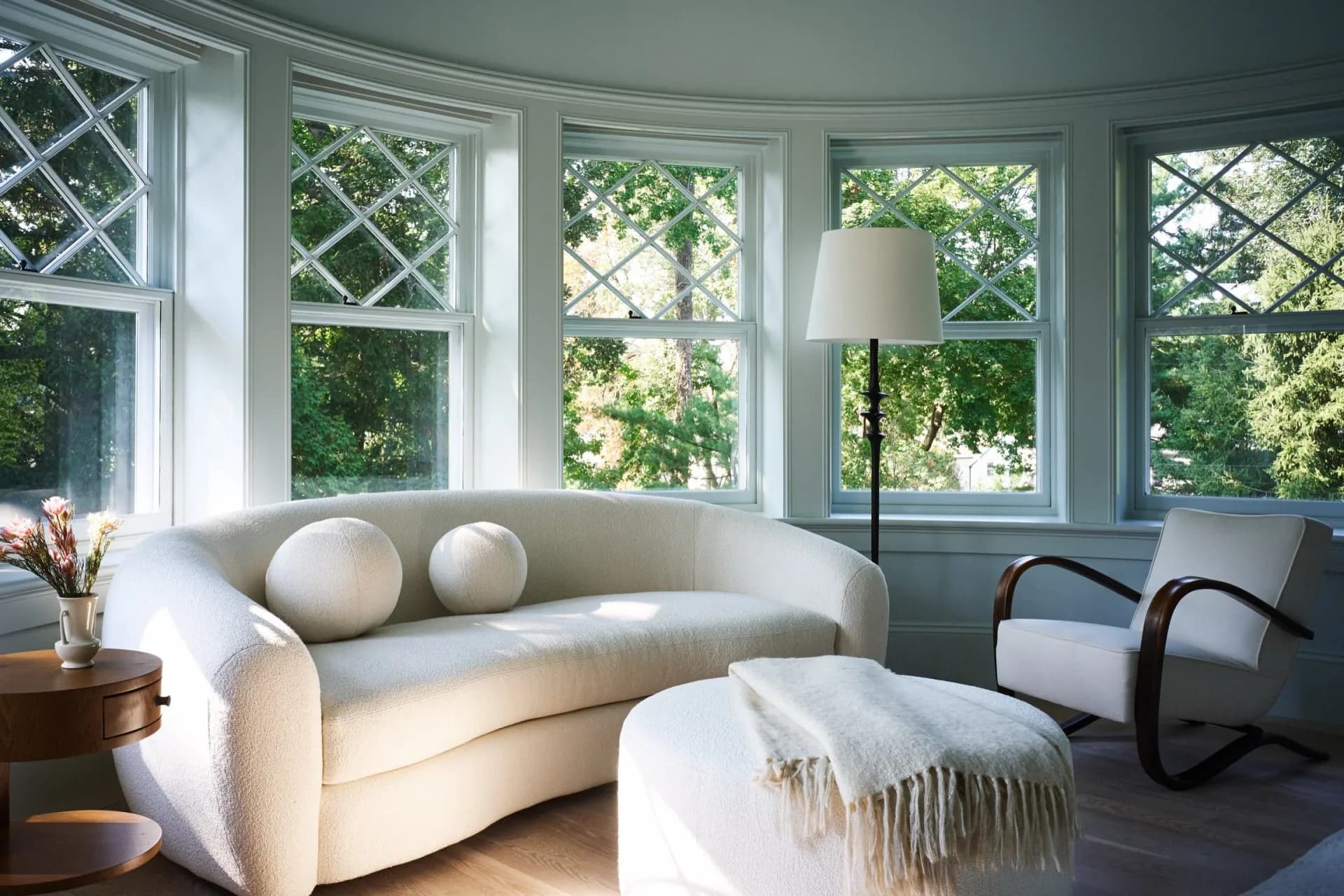 Sunlit sitting room featuring a curved cream sofa, sculptural round pillows, modern curved armchair, and ottoman with throw blanket, anchored by bay windows with geometric patterned frames overlooking green landscape, by Tusy Culos.