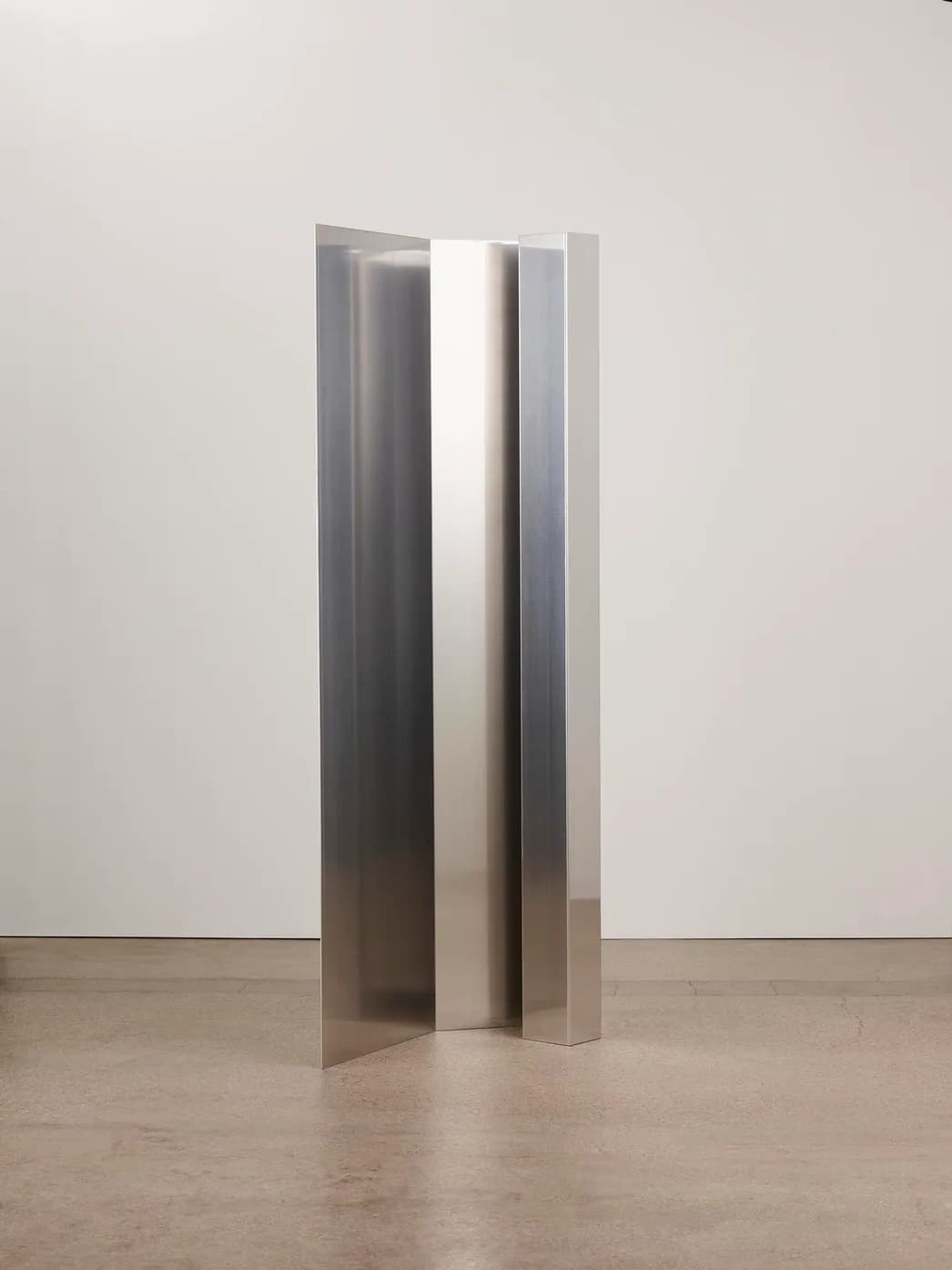 Tall sculptural metal installation with curved reflective panels in brushed steel, creating dynamic light play against white gallery walls, by Umberto Bellardi Ricci.