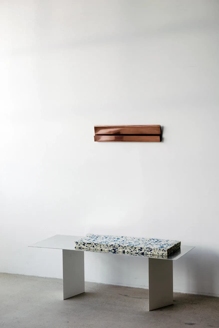 Minimalist installation featuring a concrete table with terrazzo top and glass extension, beneath a copper wall-mounted sculptural element, by Umberto Bellardi Ricci.
