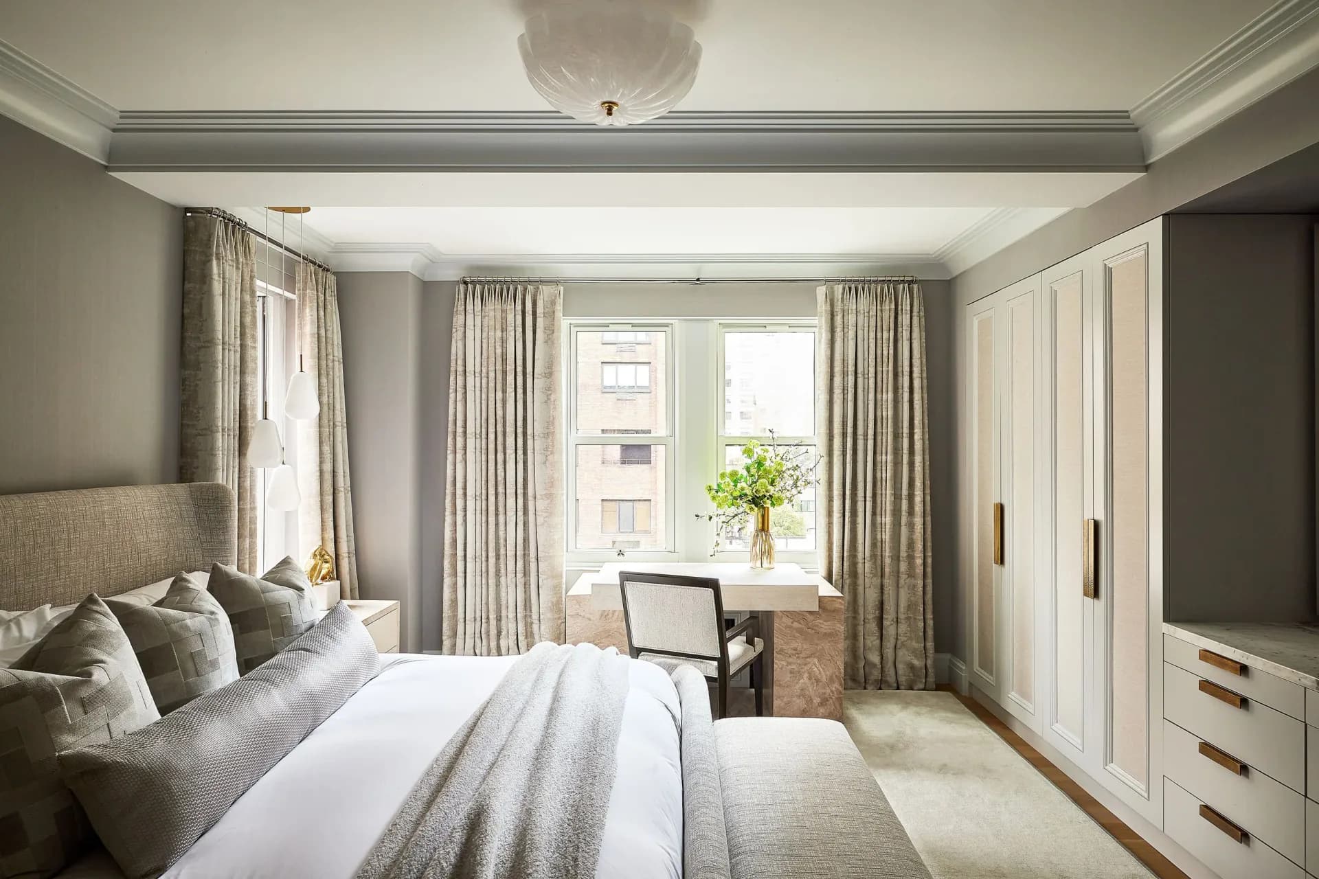 Serene bedroom with gray upholstered headboard, layered cream and taupe bedding, floor-to-ceiling windows with neutral curtains, custom white cabinetry, and coffered ceiling detail by Valerie Goldin-Remm.