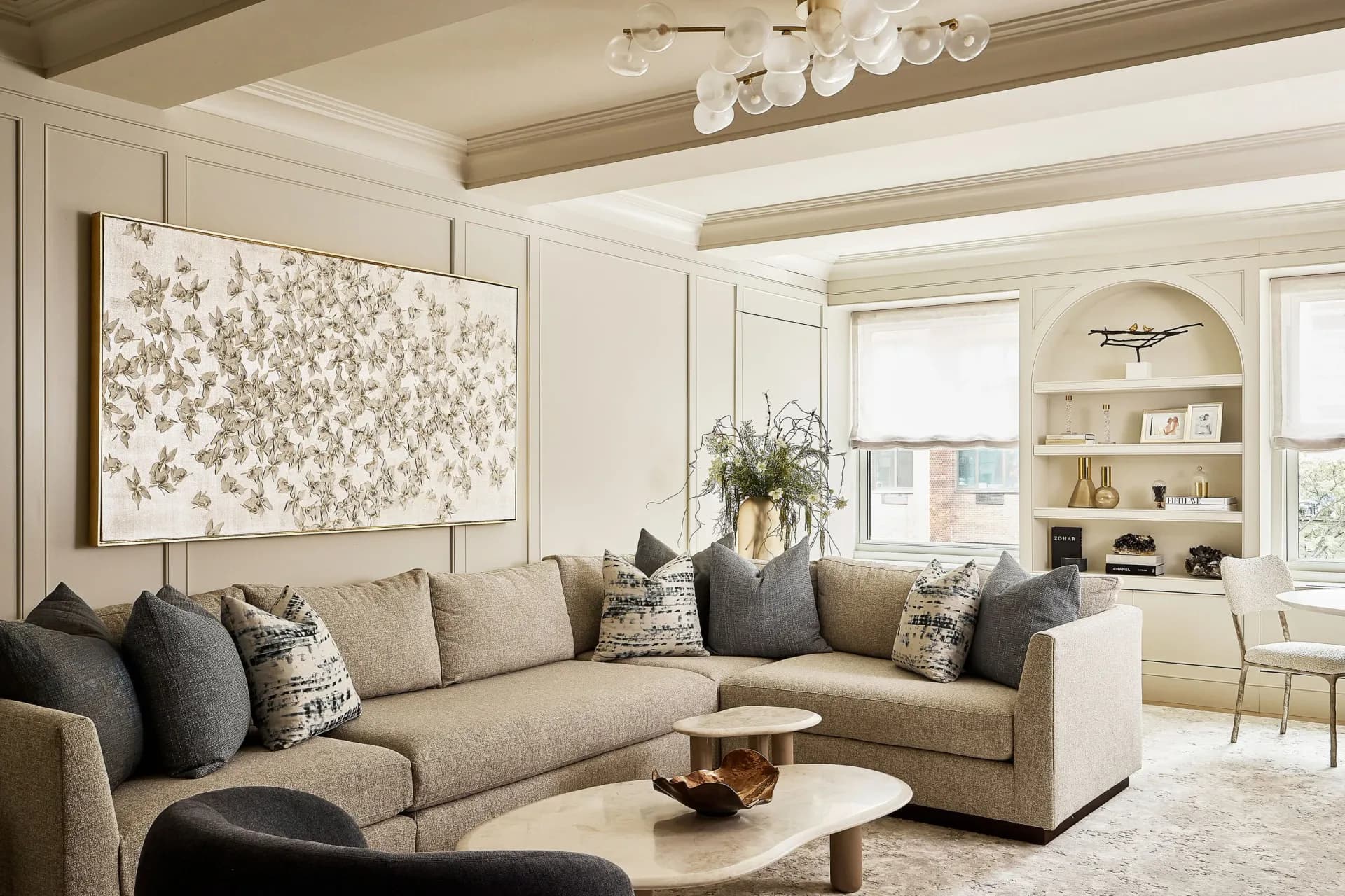 A cream and taupe living room with a sectional sofa, round marble coffee table, framed botanical artwork, and built-in shelving by Valerie Goldin-Remm.