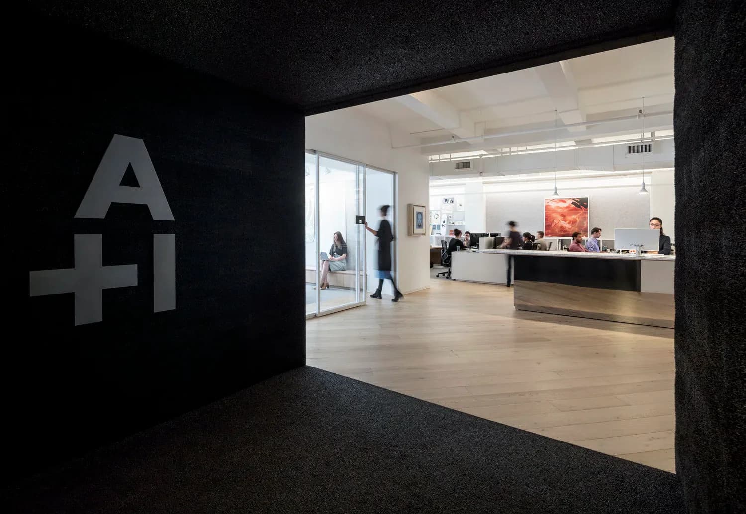 Modern office reception area with dark charcoal walls, light wood flooring, and glass partitions featuring an A+I logo, designed by Vané Broussard.