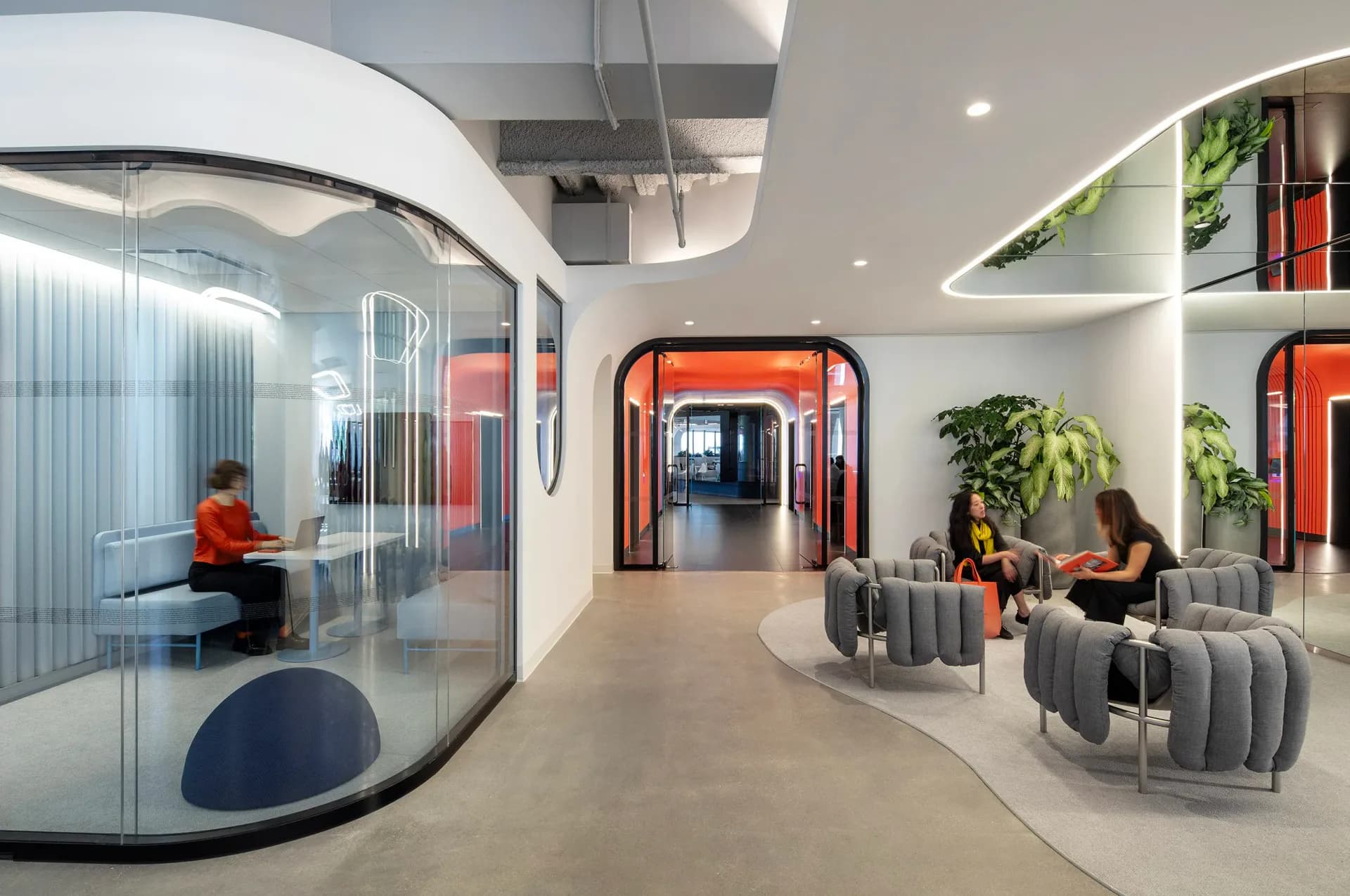 Modern office lounge with curved glass partitions, gray modular seating, orange accent walls, and living plants, designed by Vané Broussard.