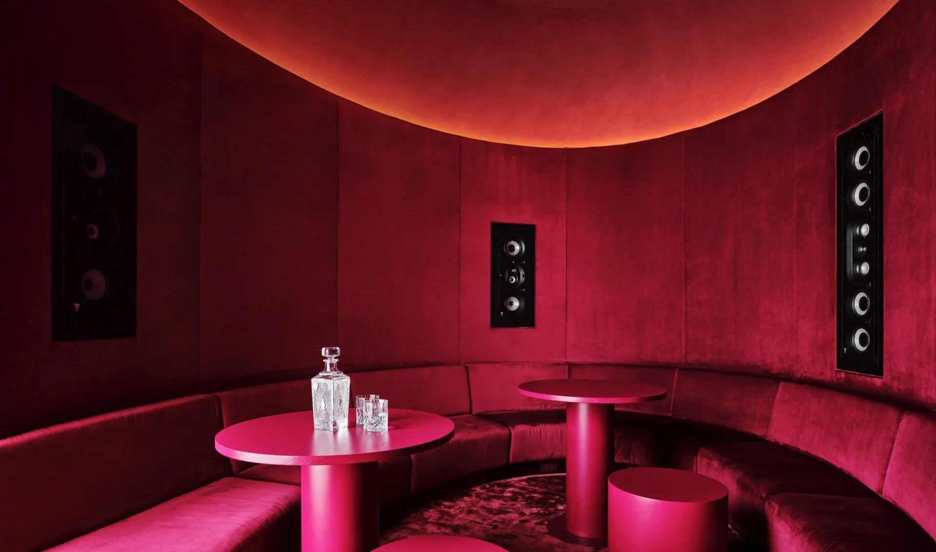 A curved lounge space with vivid magenta and red lighting, cylindrical furniture, and tall speaker columns, designed by Vané Broussard.