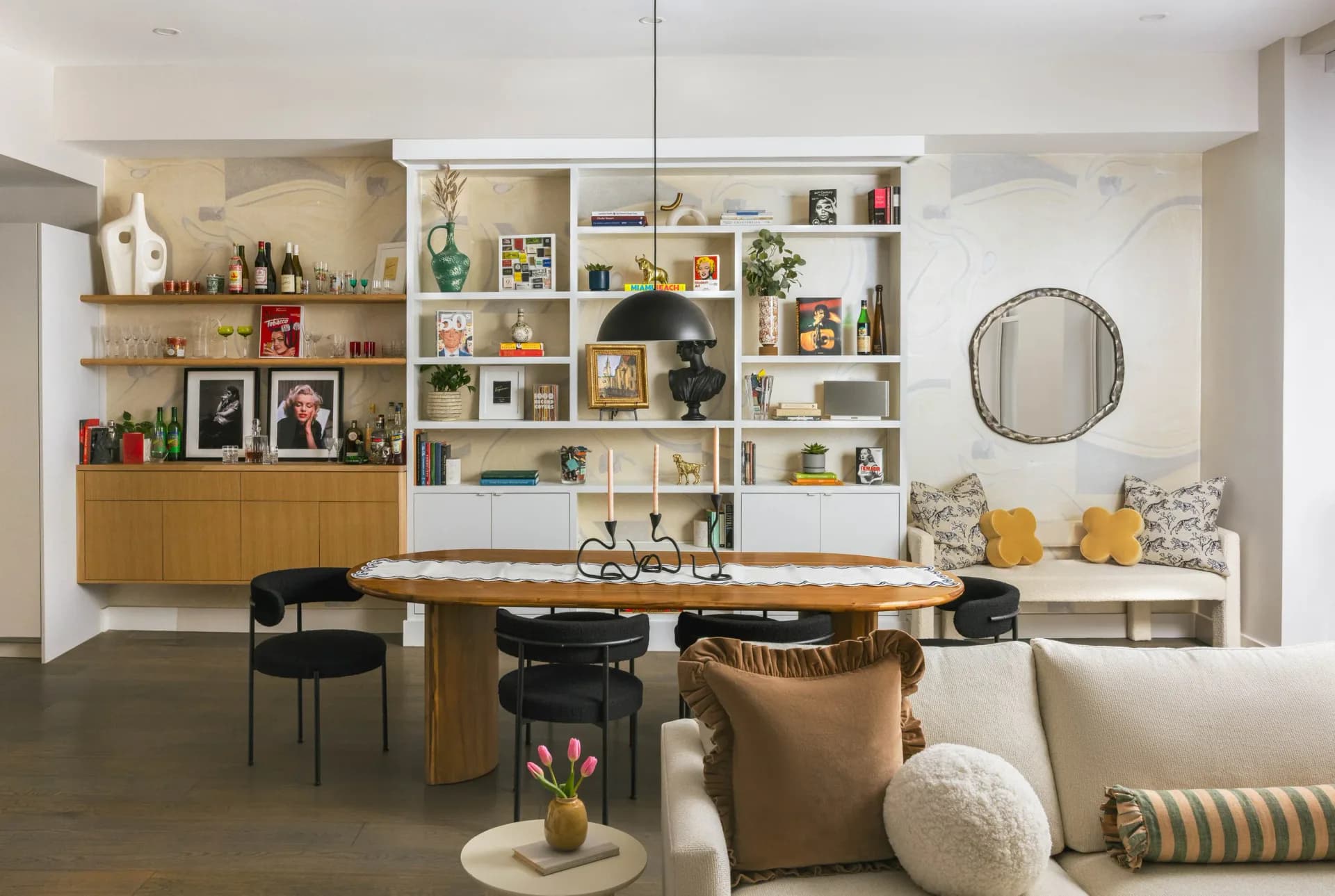 Modern living room with wood dining table, built-in shelving with warm-toned accessories, cream seating, and circular mirror by Vanessa Lugo.