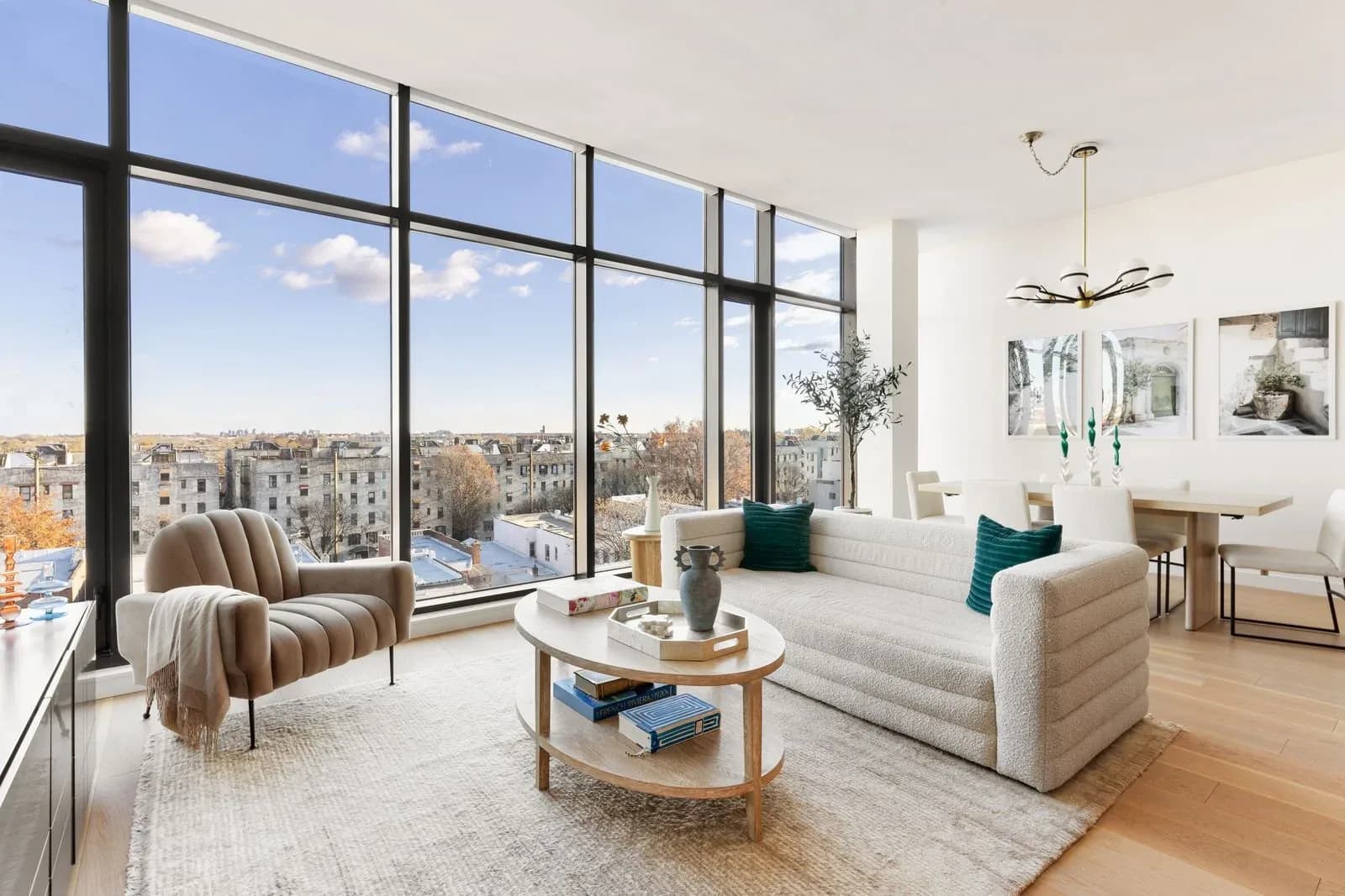Modern high-rise living room with floor-to-ceiling windows overlooking a city skyline, featuring a cream sectional sofa, striped accent chair, circular wooden coffee table, and light wood flooring by Vanessa Lugo.