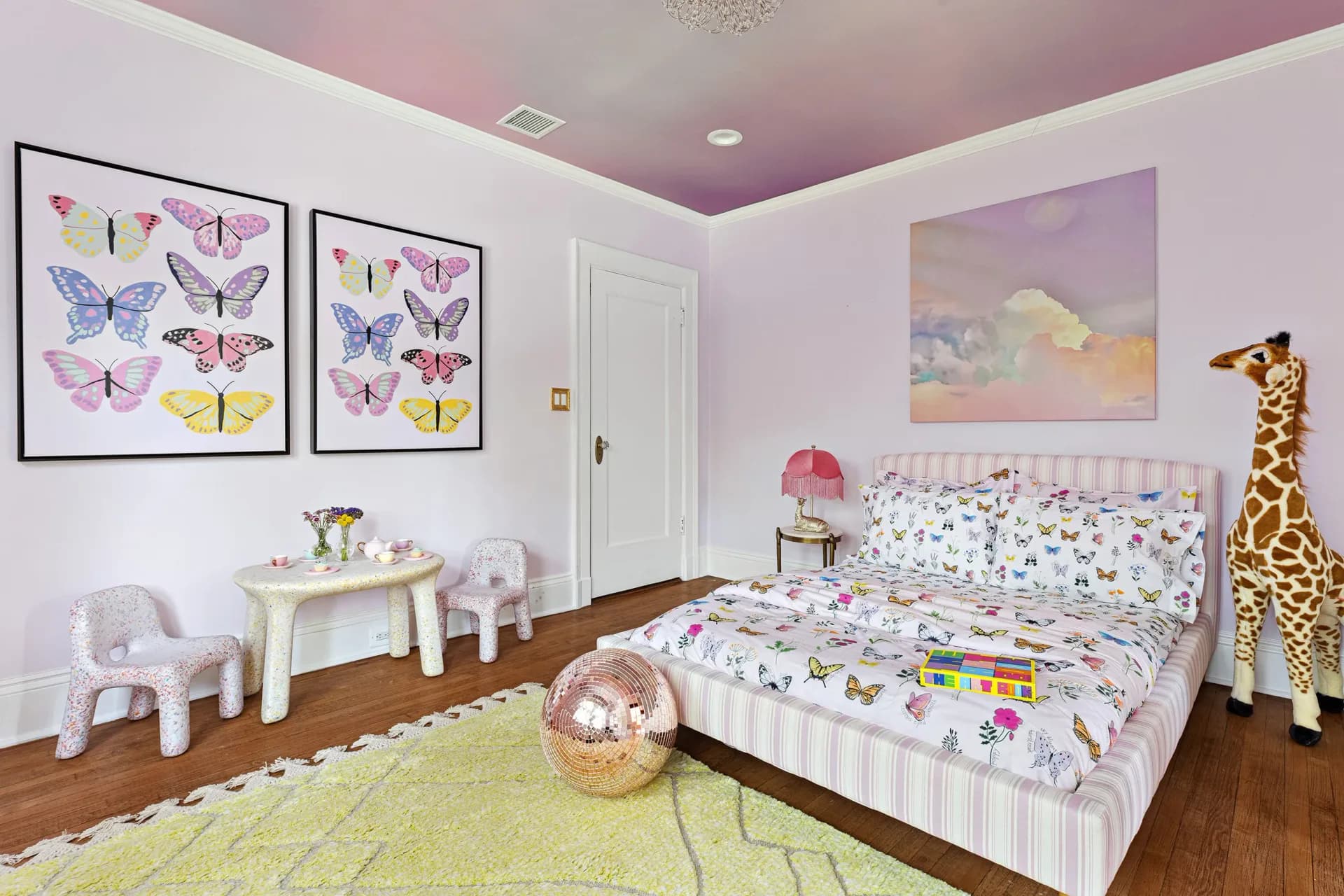 A lavender and pink children's bedroom with butterfly artwork, white furniture, floral bedding, and a wooden giraffe toy, designed by Vanessa Lugo.