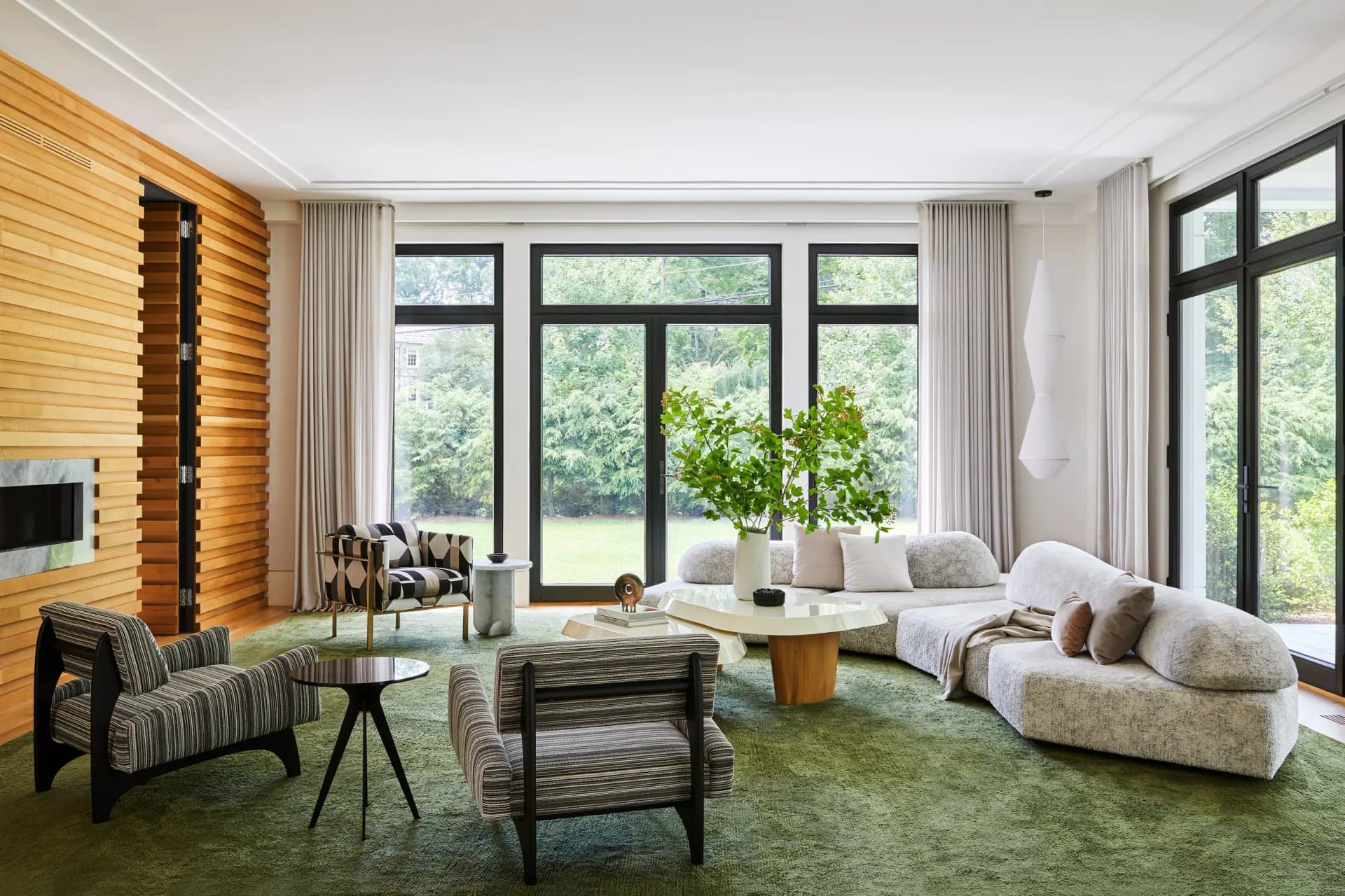 Modern living room with natural wood accent wall, curved cream furniture, green area rug, and expansive windows overlooking greenery by Vanessa Rome.